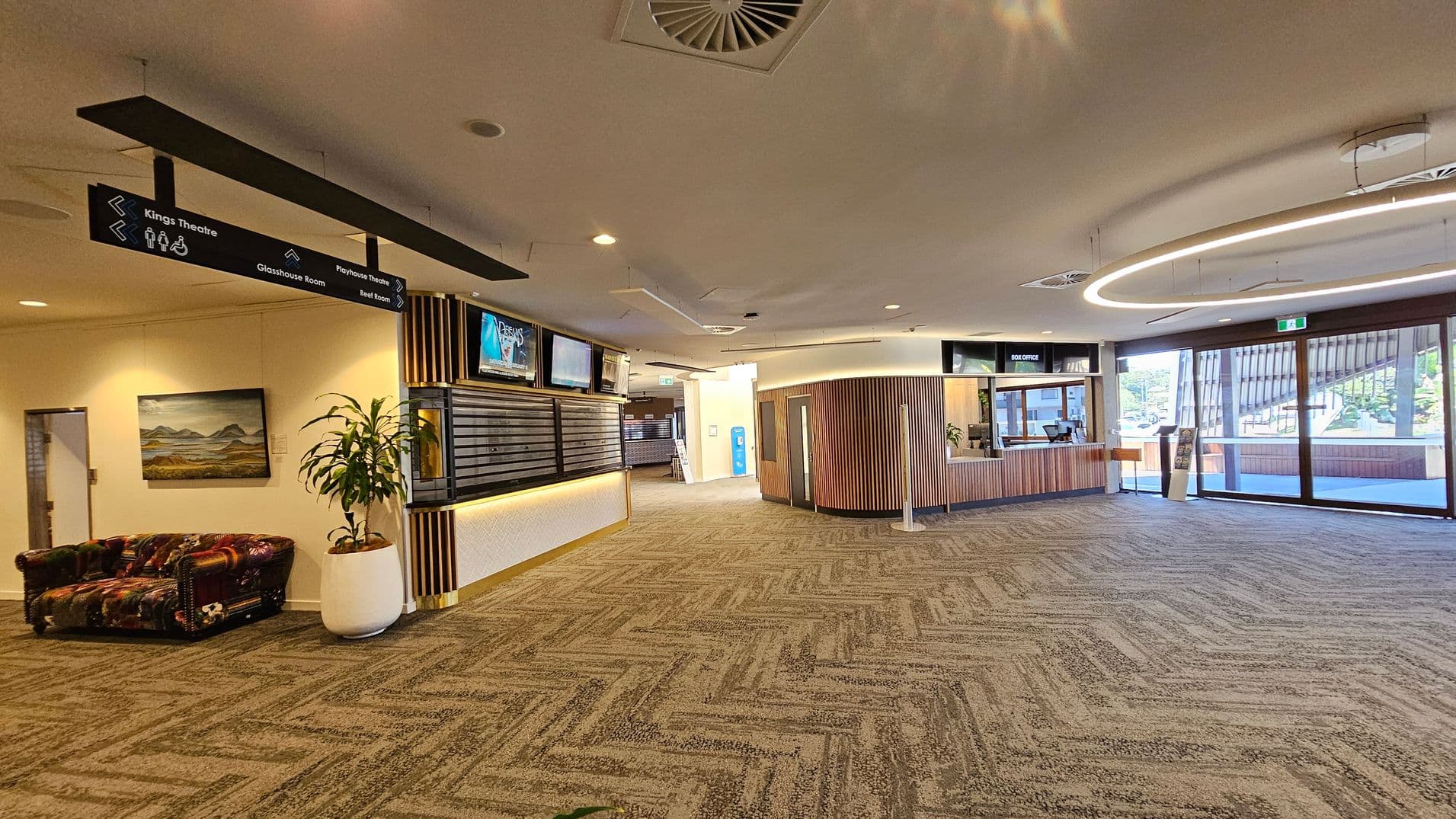 Spacious lobby with patterned carpet flooring. On the left, a colourful sofa and a painting on the wall. Overhead signs direct to various rooms. In the centre, a wooden reception desk with digital screens above. A large potted plant is nearby. On the right, a box office counter with a circular light fixture above. Glass doors in the background lead outside, allowing natural light to enter. The ceiling is white with recessed lighting and air vents.