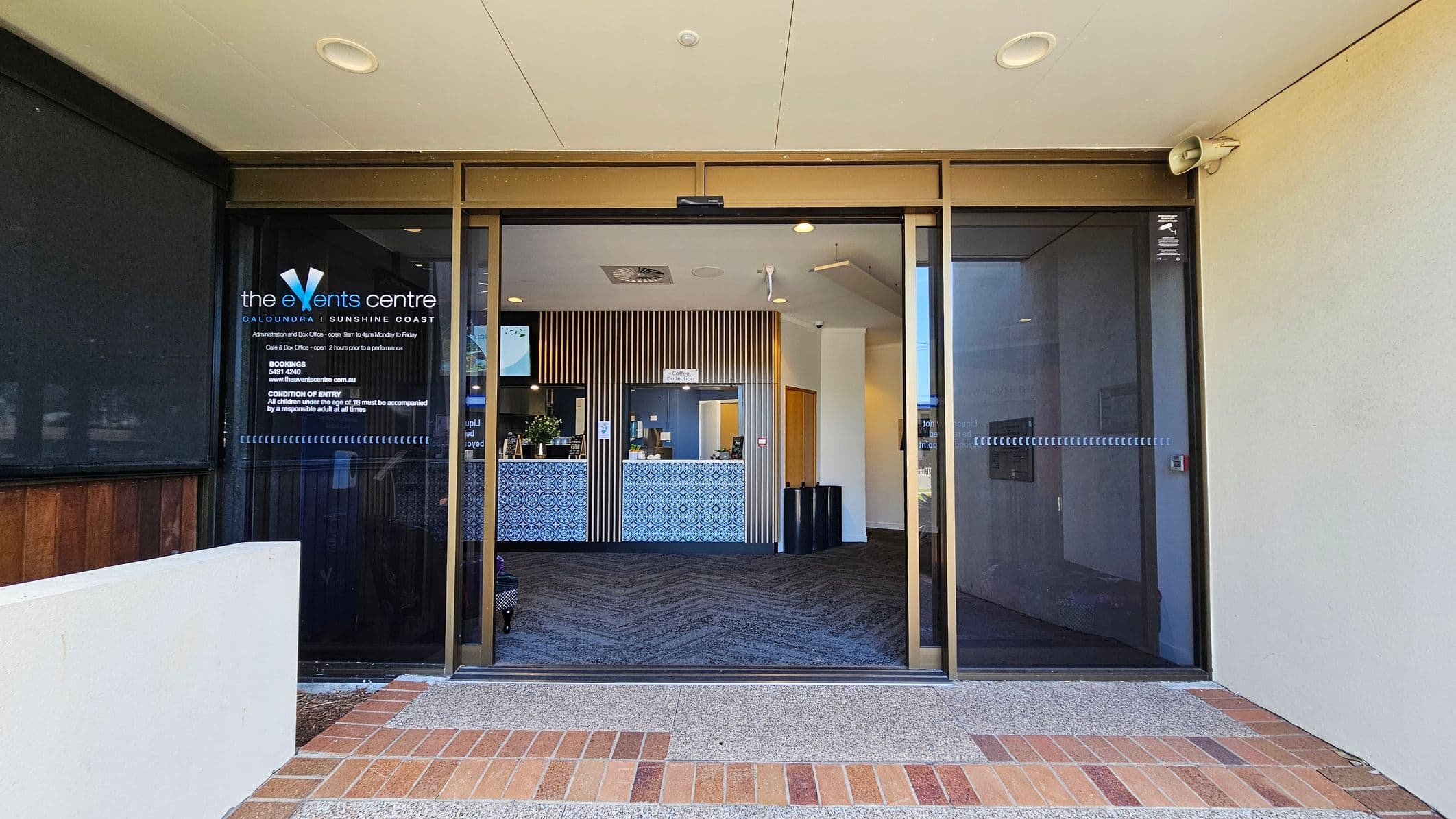 Entrance to a building with glass sliding doors. On the left, there's a sign for "the Events Centre" with details about opening hours and conditions of entry. Inside, a reception area with a patterned counter and vertical wooden slats on the wall. The floor is carpeted with a zigzag pattern. The ceiling is light-coloured with recessed lighting. The exterior floor is a mix of brick and concrete.