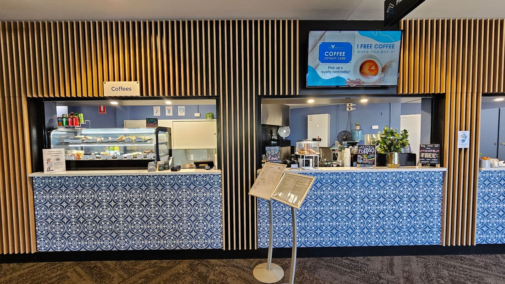 A café counter with two service windows, each adorned with blue and white patterned tiles. The left window displays a variety of snacks and drinks in a glass case, with a sign above reading "Coffees". The right window features a coffee machine and a small plant. Above, a digital screen advertises a coffee loyalty card. The background has vertical wooden slats, and a menu stand is positioned in front of the counter. The floor is carpeted with a geometric pattern.