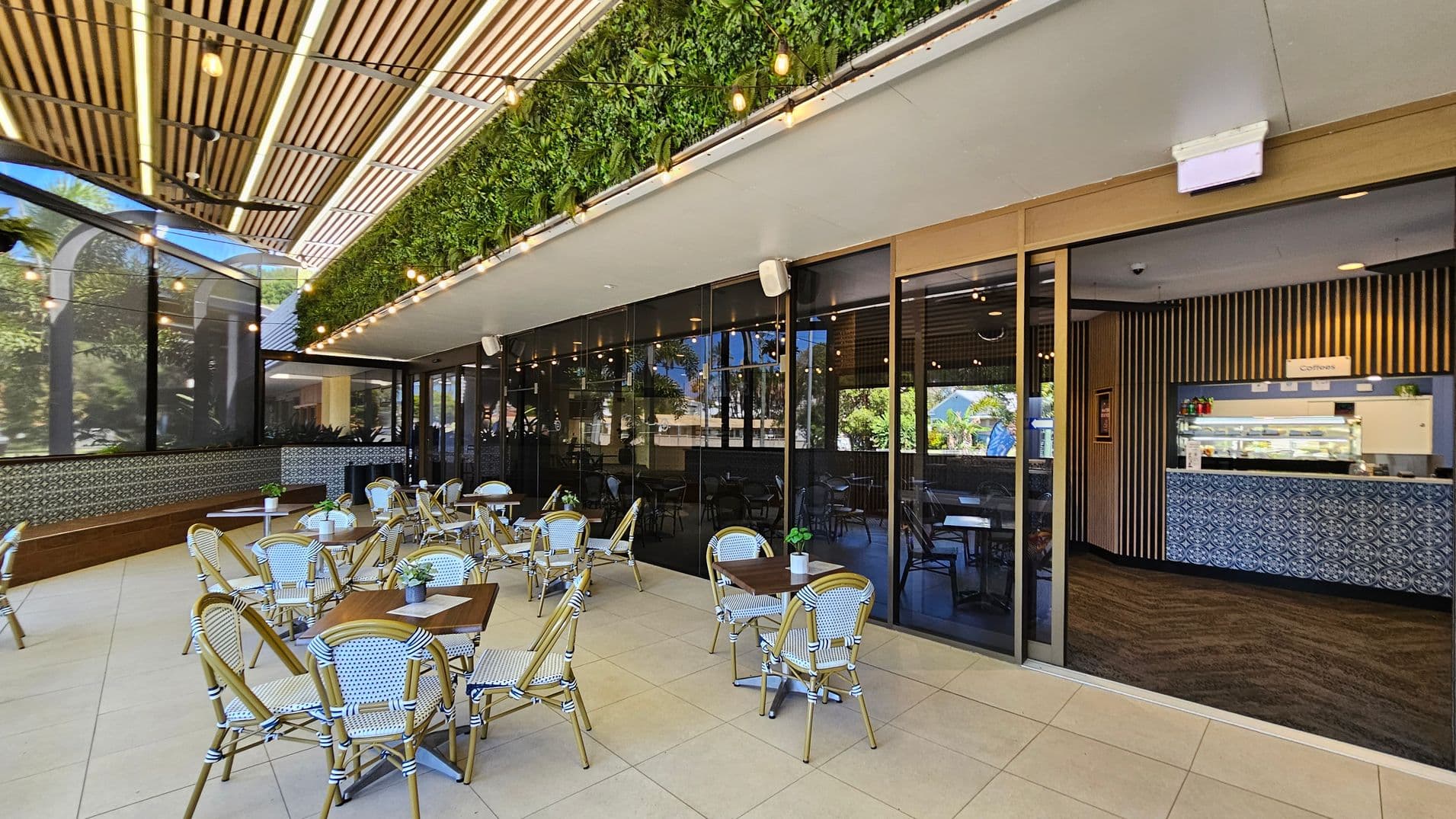 Outdoor café area with several tables and chairs on a tiled floor. On the left, a wooden bench runs along a wall with decorative tiles. The ceiling features wooden slats and string lights. A vertical garden with lush greenery is above the glass wall on the right, which separates the outdoor seating from the indoor café. Inside, a counter with patterned tiles displays a sign for coffee. Large windows reflect the surrounding greenery.