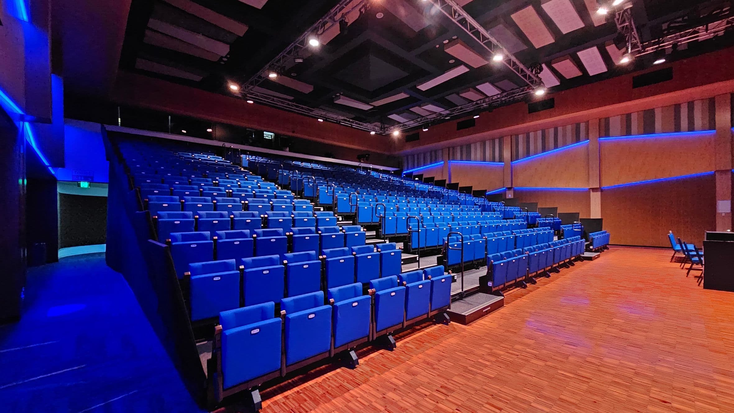 An auditorium with rows of blue seats arranged in tiers on the left and centre. The floor is wooden with a warm tone. The walls are lined with vertical panels and illuminated by blue LED lights. The ceiling features a grid of acoustic panels and spotlights. On the right, there is a small area with additional seating and a table.