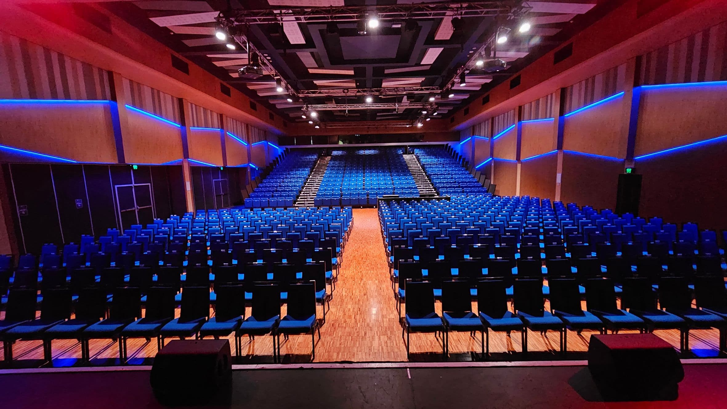 A large auditorium with rows of blue chairs arranged in a semi-circular pattern. The walls are lined with blue LED lights, creating a modern look. The ceiling features a geometric design with spotlights. The floor is wooden, and there are aisles dividing the seating sections. The view is from the stage, looking towards the back of the auditorium.