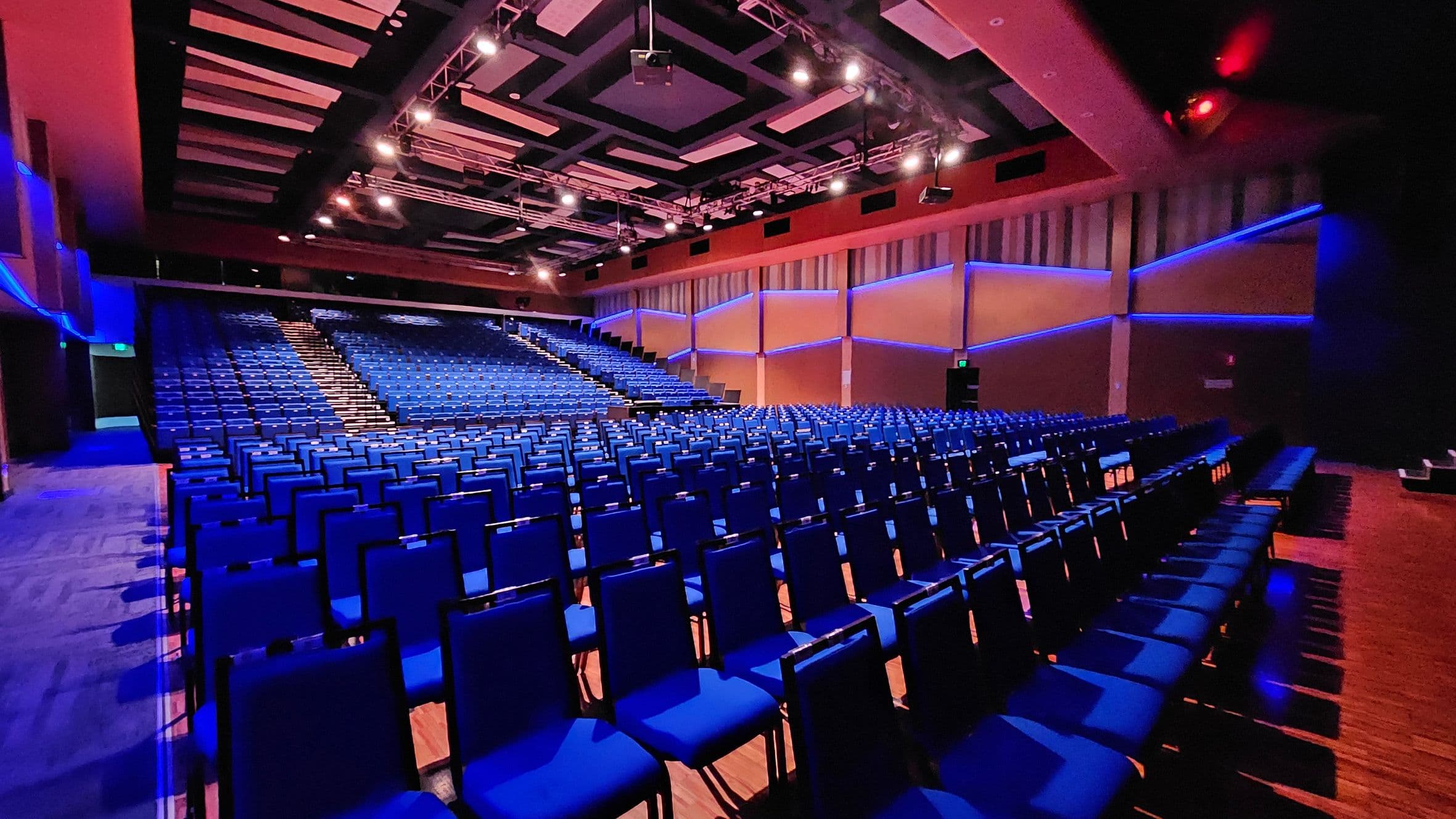 Rows of blue chairs fill a large auditorium, with a central aisle leading up to a stage. The walls are adorned with geometric patterns and blue lighting accents. The ceiling features a complex grid of lights and panels. The floor is wooden, and the space is well-lit, creating a vibrant atmosphere.