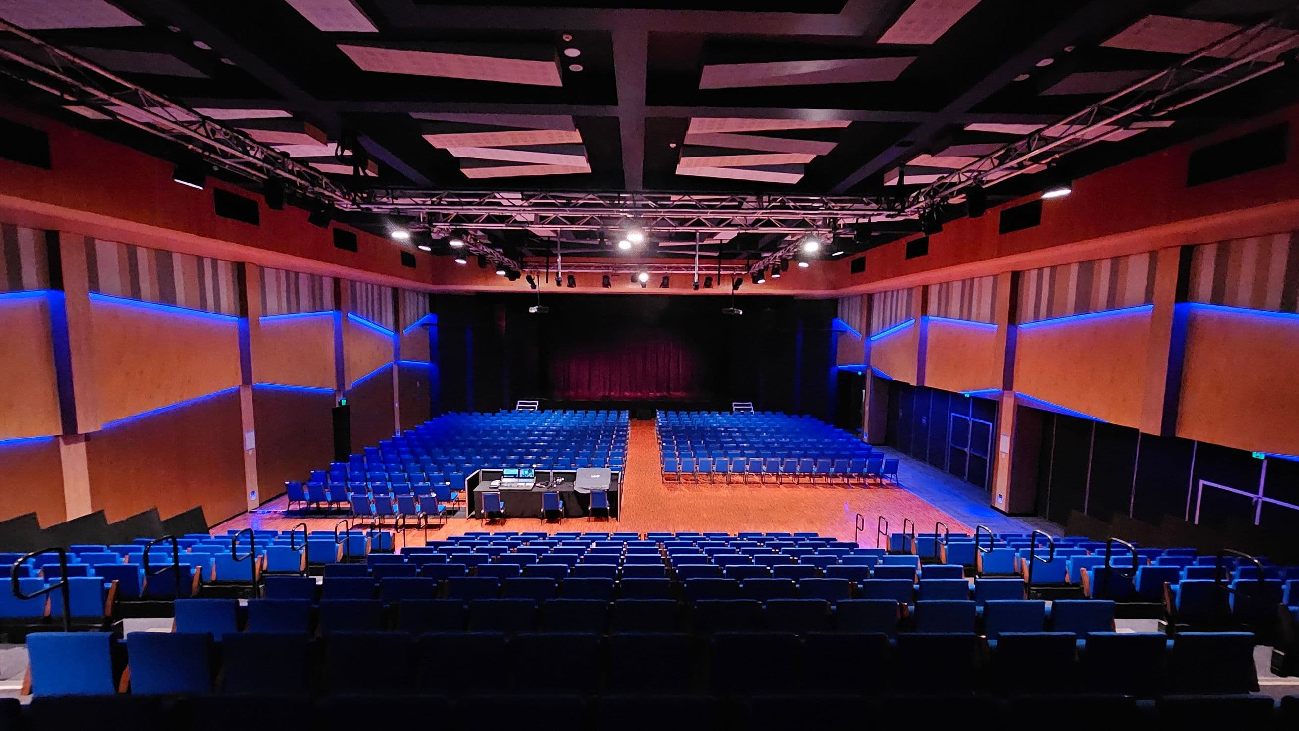 A large auditorium with rows of blue seats facing a stage at the back. The stage is framed by dark curtains. In the centre, there is a control desk with equipment and chairs. The walls are lined with vertical panels and illuminated by blue lights. The ceiling features a grid of beams and spotlights. The floor is wooden, and there are handrails along the aisles.