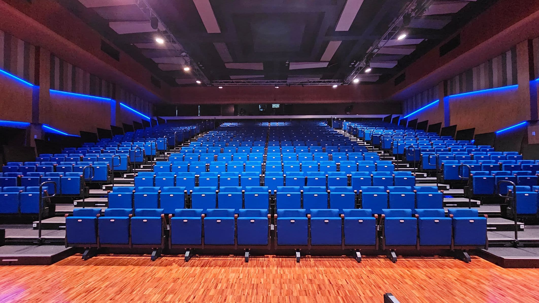 Rows of blue theatre seats are arranged in a large auditorium. The seats are on a sloped floor, leading up to the back of the room. The walls are lined with blue LED lights, creating a modern atmosphere. The ceiling is equipped with rectangular panels and spotlights. The floor is wooden, adding warmth to the space.