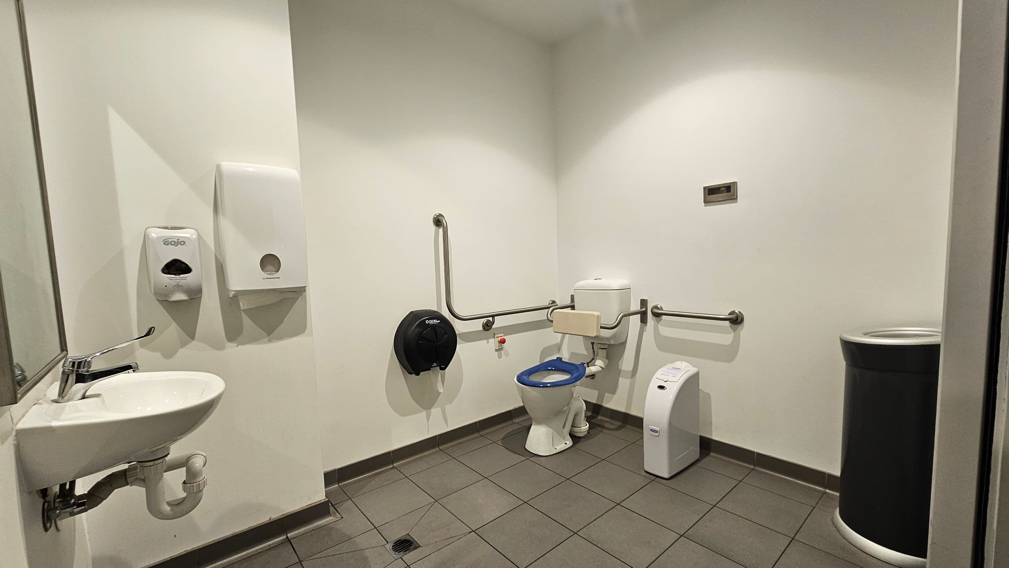 Accessible bathroom with a white sink and a mirror on the left wall. Above the sink are a soap dispenser and a paper towel dispenser. In the centre, a toilet with a blue seat is equipped with support rails on both sides. A black toilet paper dispenser is mounted on the wall beside it. On the right, a sanitary bin is placed against the wall. The floor is tiled in a light brown colour, and the walls are painted white.
