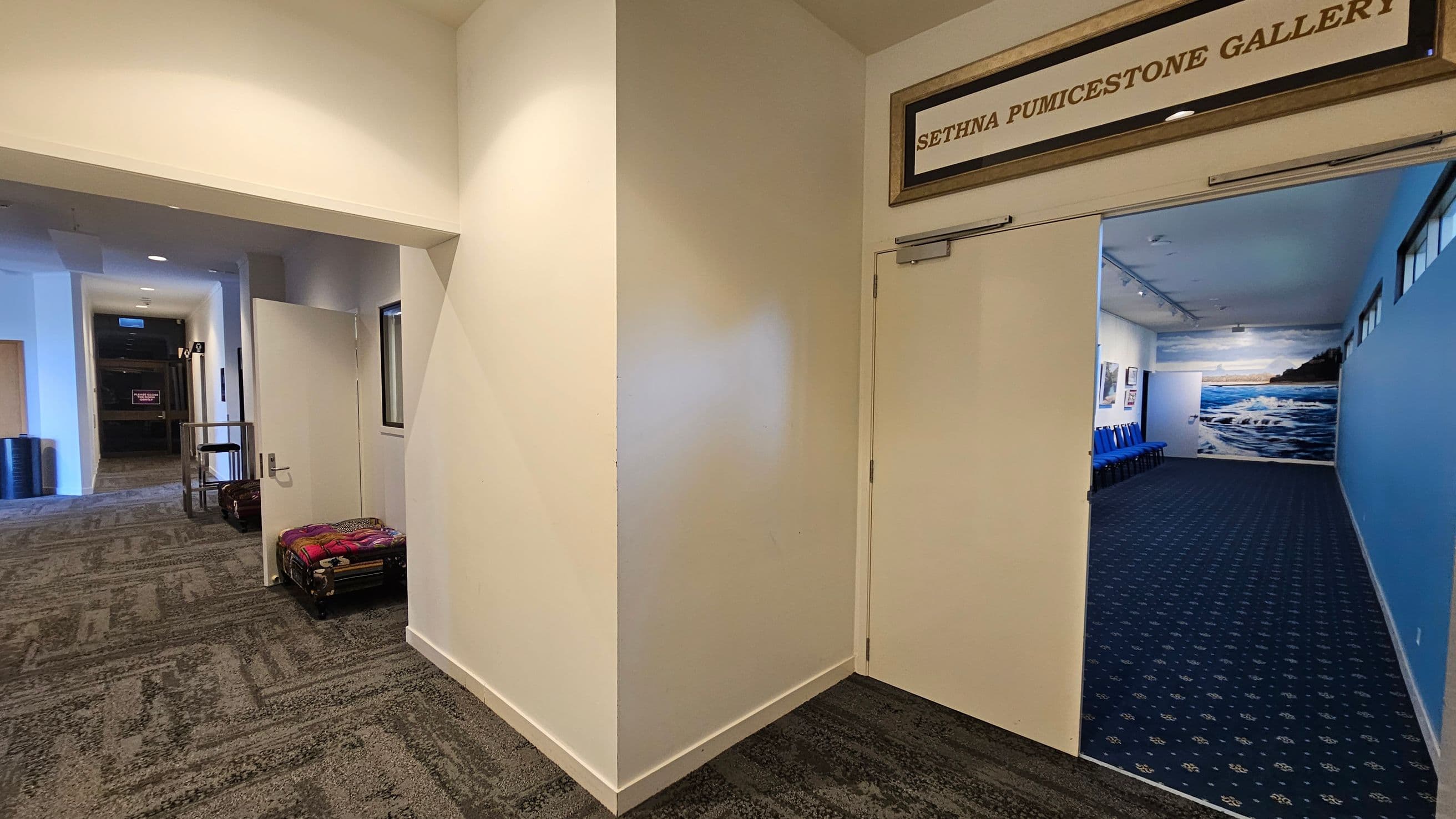 A hallway with a patterned carpet leads to an open door on the right, marked "Sethna Pumicestone Gallery." Inside, the gallery has blue walls and a mural of a seascape. Blue chairs are lined up against the left wall. On the left side of the hallway, there is a partially open door and a trolley with luggage. The ceiling has recessed lighting, and the walls are painted in a light colour.