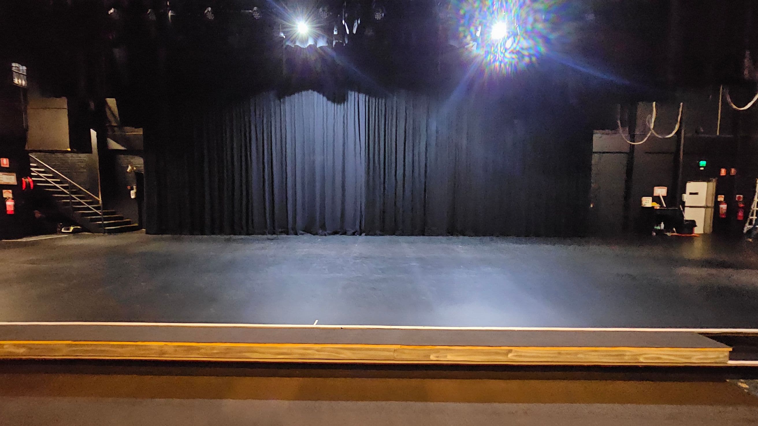 A theatre stage with a black curtain backdrop. The floor is dark and smooth, extending to the front of the stage. On the left, there is a staircase leading up to a higher level. On the right, there are some technical equipment and a ladder. Overhead, stage lights are visible, casting a bright glow. The ceiling is not visible.
