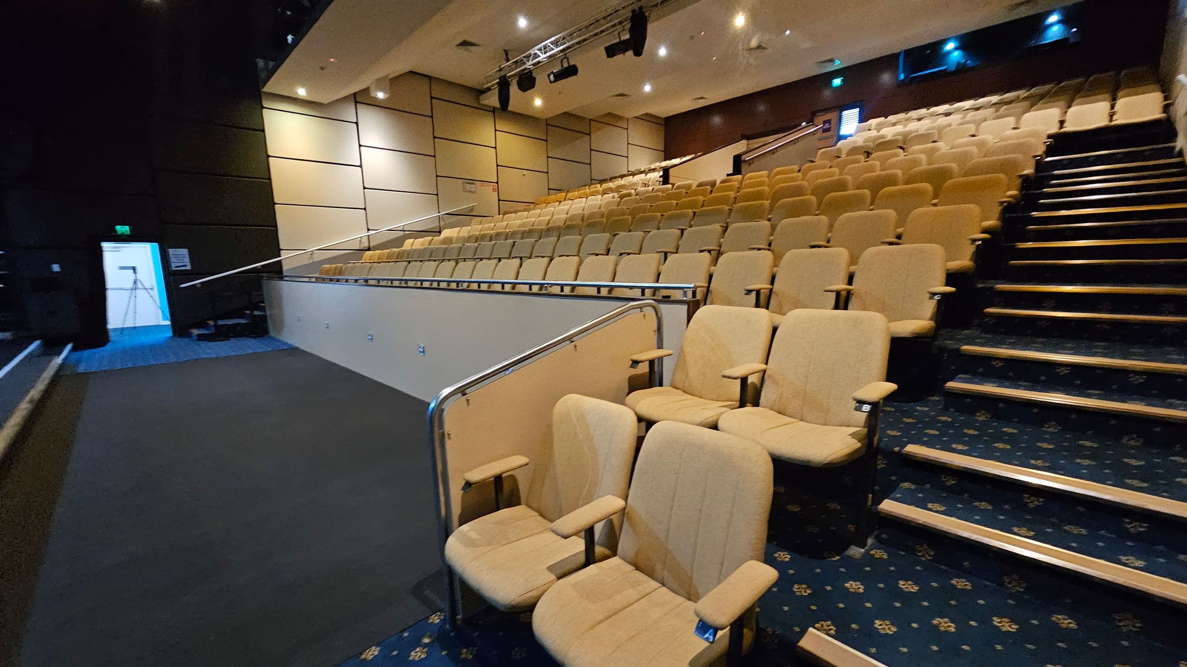 A lecture hall with tiered seating on the right, featuring beige upholstered chairs. The floor is carpeted in a dark blue pattern. On the left, there is a walkway with a metal railing and a door with a blue light visible through it. The walls are panelled in a light colour, and the ceiling has recessed lighting and mounted speakers.