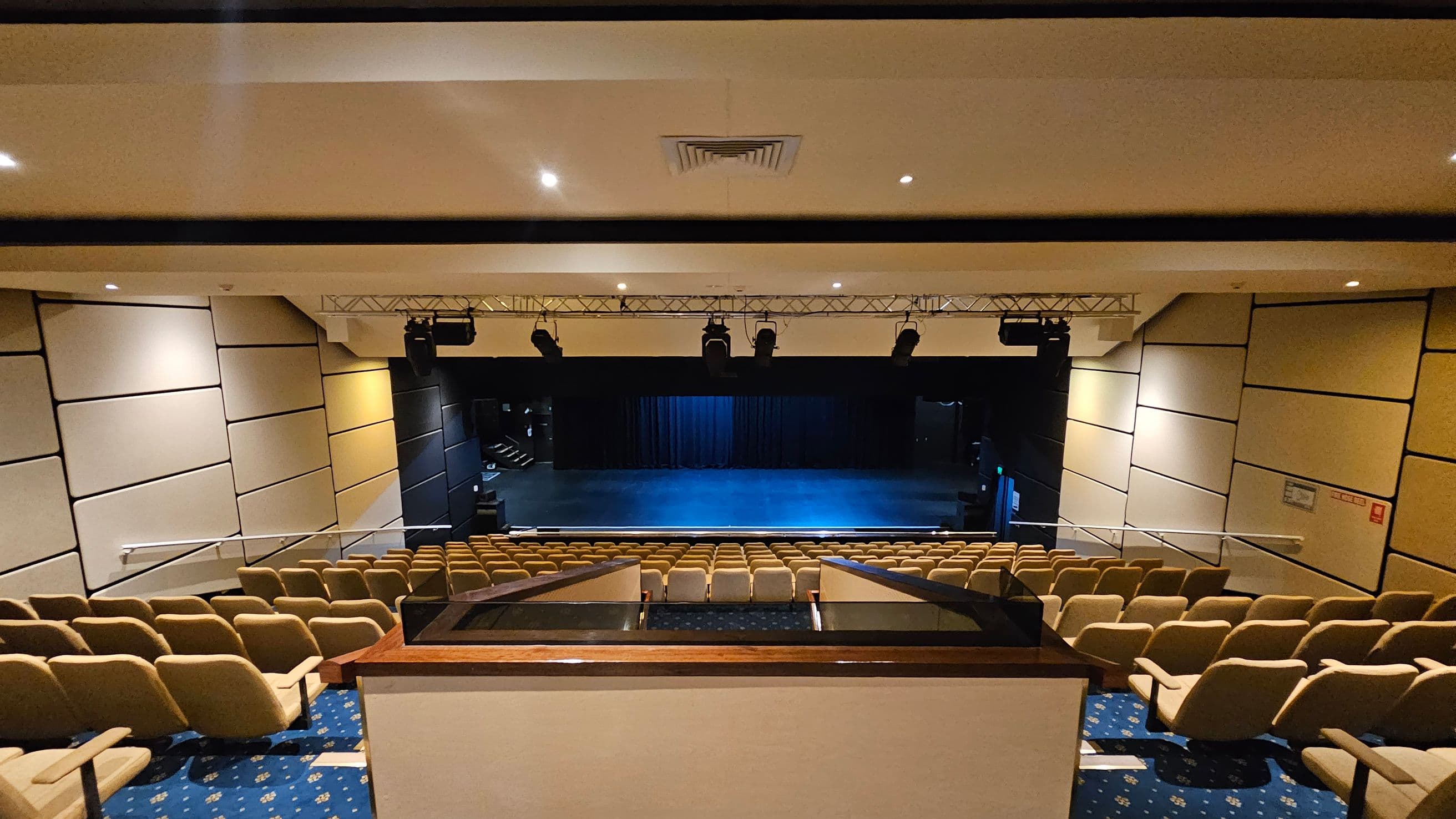 A theatre interior with rows of beige seats facing a stage. The stage is empty with a blue-lit floor and a black curtain backdrop. Overhead, there are stage lights mounted on a metal truss. The walls are lined with beige acoustic panels. The floor is carpeted in blue with a pattern, and the ceiling is white with recessed lighting and a central air vent.