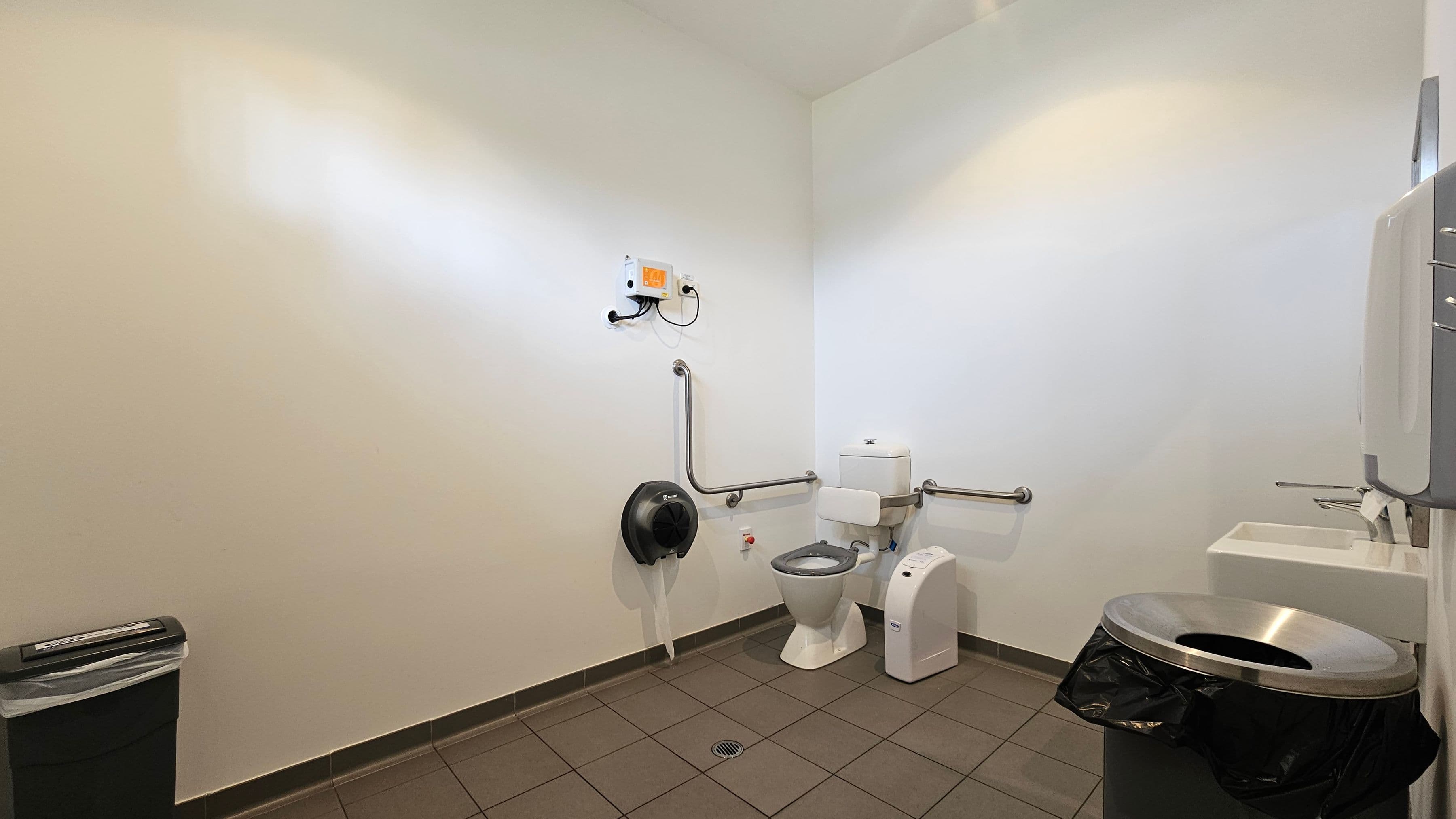 A public restroom with a white tiled floor and white walls. On the right, there's a sink with a soap dispenser above it. Next to the sink is a toilet with support rails on both sides. Above the toilet, a small white box is mounted on the wall. To the left of the toilet, a large toilet paper dispenser is attached to the wall. In the foreground on the left, a black bin with a silver lid is visible.