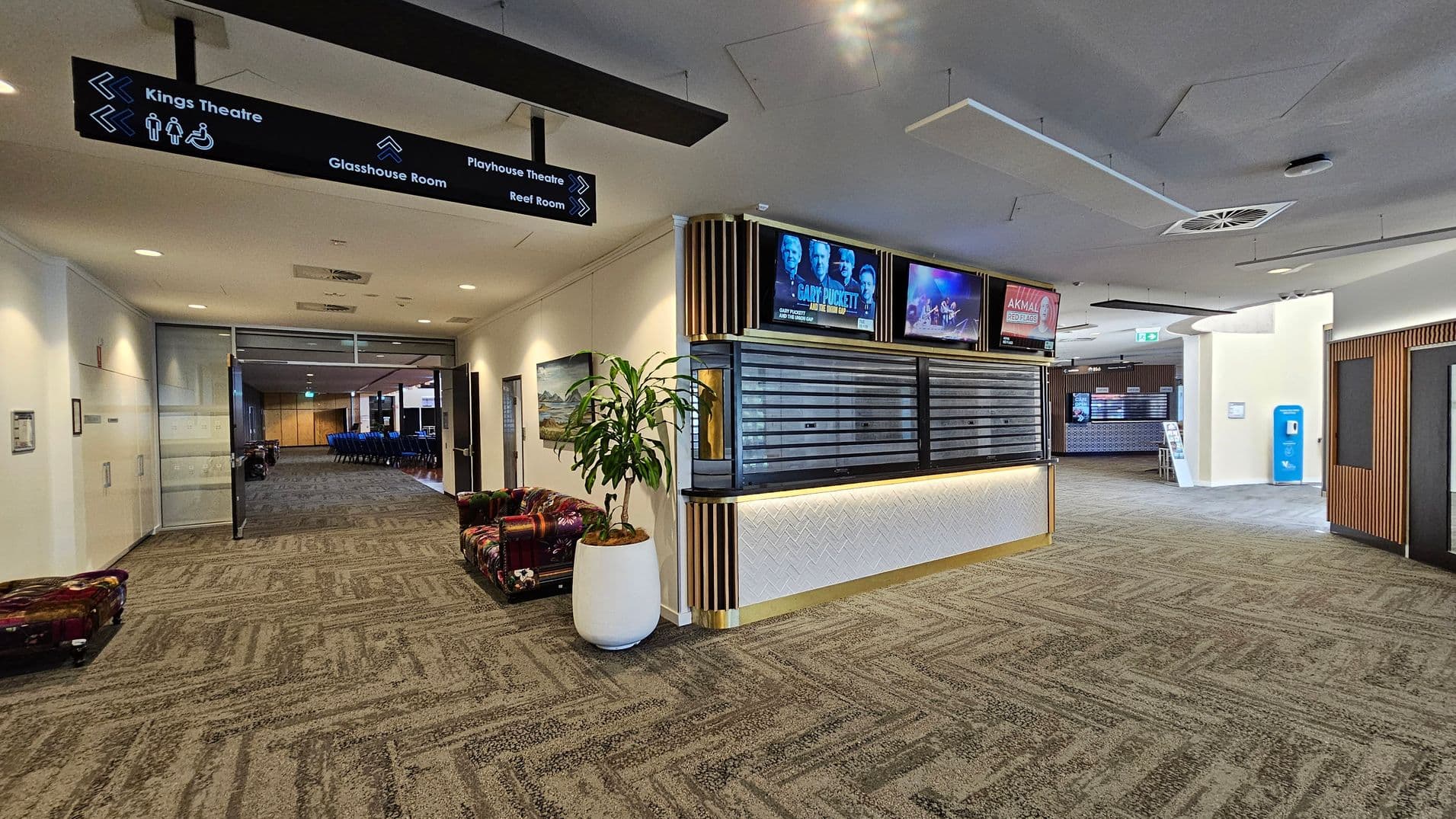 A spacious lobby area with a patterned carpet floor. On the left, there is a hallway leading to a room with blue chairs, and a sign above pointing to various locations like Kings Theatre and Glasshouse Room. A potted plant sits near a colourful cushioned bench. In the centre, a counter with digital screens above displays event information. On the right, a blue vending machine is visible near another hallway. The ceiling has recessed lighting and air vents.