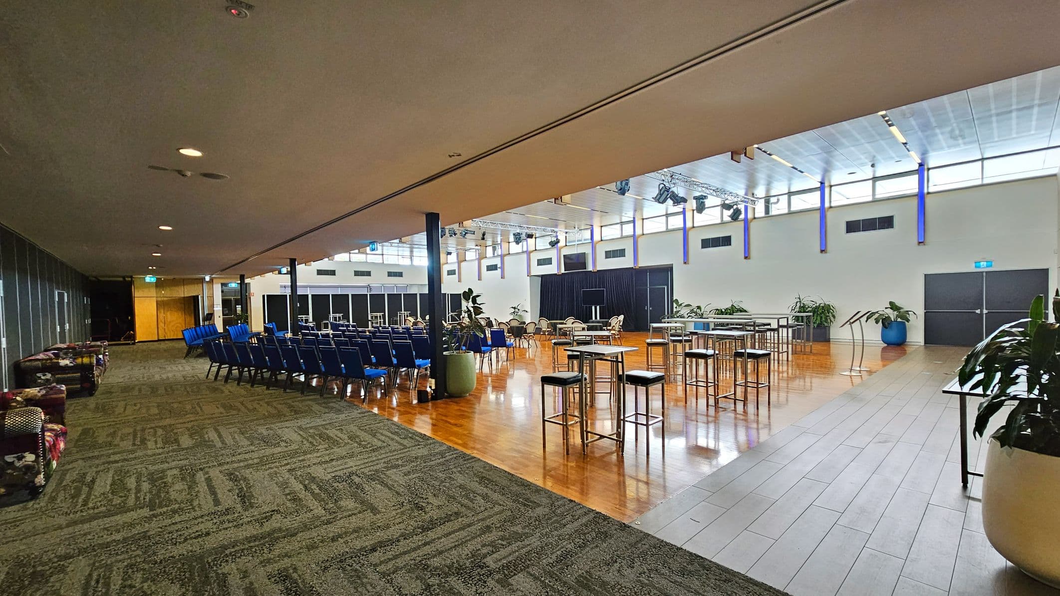 A spacious indoor venue with a mix of carpeted and wooden flooring. On the left, there are rows of blue chairs facing a stage area. The right side features high tables with stools and potted plants. The ceiling is high with large windows allowing natural light. The walls are white with blue vertical accents, and there are doors at the far end. The ceiling has lighting fixtures and speakers.