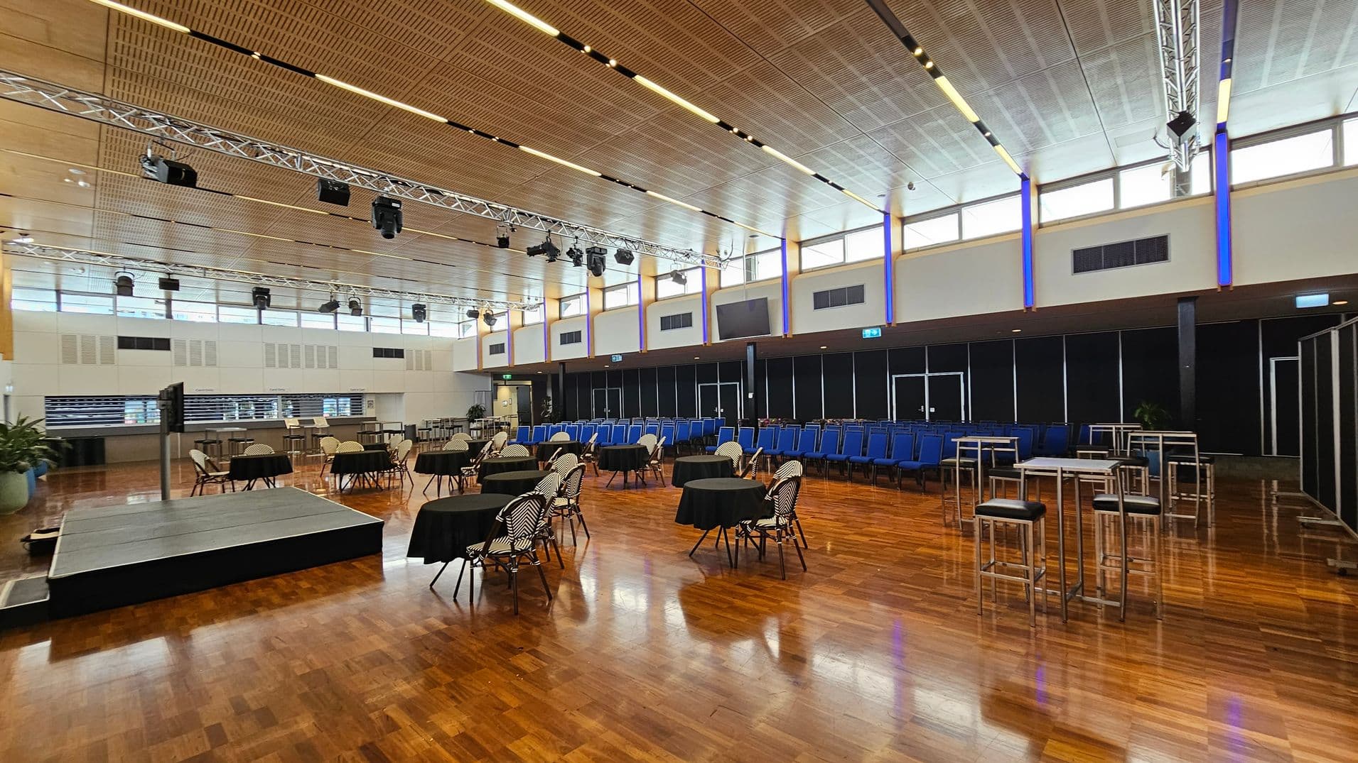 A spacious indoor venue with rows of blue chairs arranged in the centre on a wooden floor. To the left, there are tall tables and chairs, along with potted plants. The right side features a carpeted area with additional seating and tables. The ceiling is high with visible lighting fixtures, and large windows allow natural light to enter. In the background, there are more plants and a dark curtain. The far right shows an open corridor leading to another area.