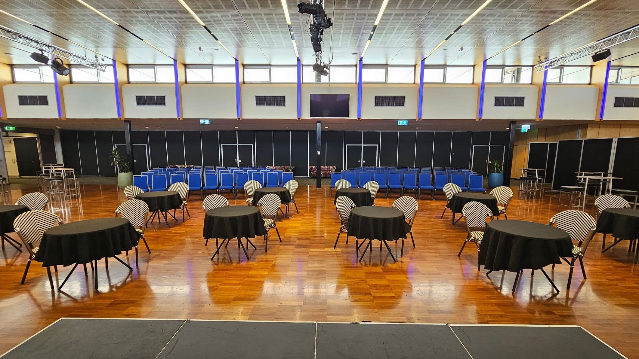 A spacious hall with a wooden floor and a high, panelled ceiling. On the left, there is a small stage and several round tables with black tablecloths and chairs. The right side features rows of blue chairs and a few tall tables with stools. The back wall has large windows and black panels, with lighting fixtures and speakers suspended from the ceiling.