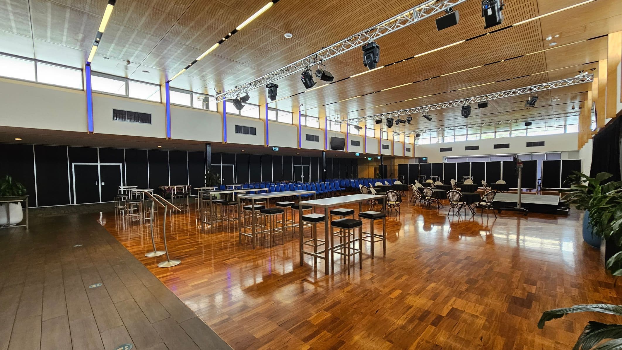 A spacious hall with a polished wooden floor. In the foreground, several round tables are covered with black tablecloths, surrounded by chairs with black and white striped covers. In the background, rows of blue chairs are neatly arranged. The ceiling is high with a grid pattern and features lighting fixtures. The walls have large windows near the ceiling, allowing natural light to enter. On the sides, there are tall plants in pots and some high tables with stools.
