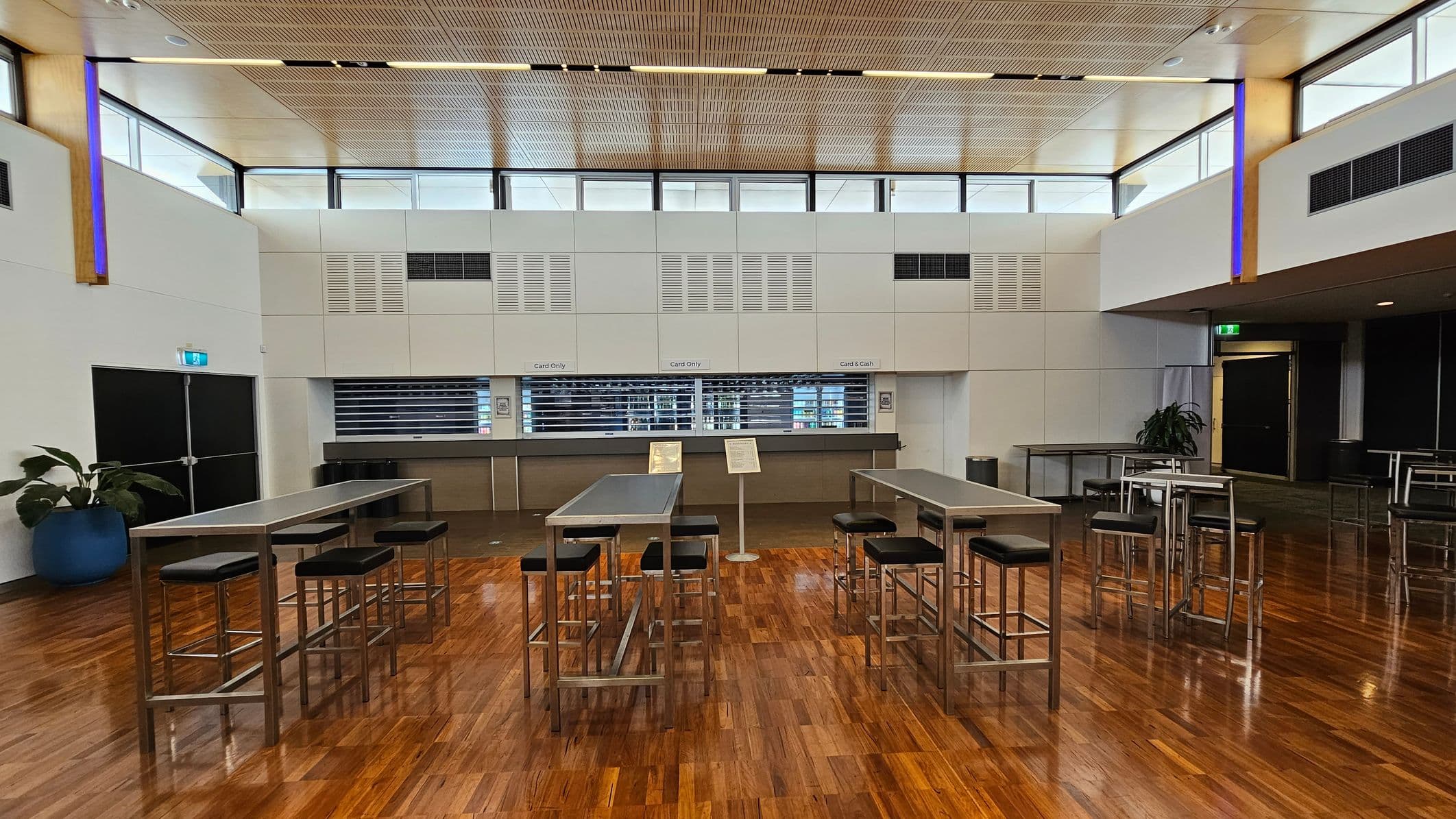 A spacious indoor venue with a polished wooden floor. On the left, there are high tables with stools and a few potted plants. In the centre, rows of blue chairs are arranged in front of a stage area. On the right, round tables with chairs are scattered. The ceiling is high with wooden panels and equipped with lighting and speakers. Large windows near the ceiling allow natural light to enter.