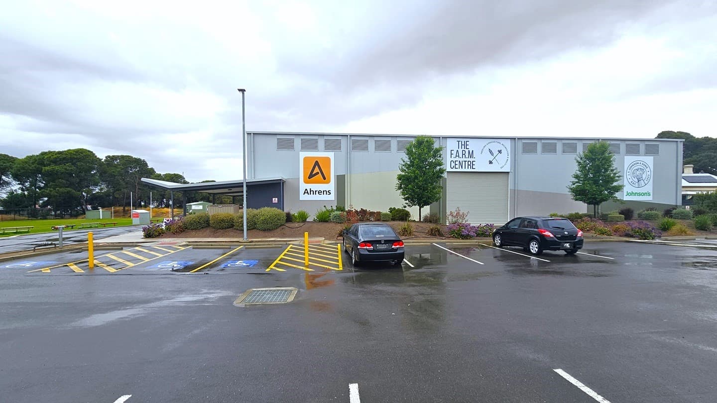 The car park is located near the building. Accessible parking is marked with blue wheelchairs, yellow diagonal lines, and bollards. Two black cars are parked. Behind the car park is a grey industrial-type building with the sign "The Farm Centre." Advert signs are displayed on the building wall. The building is surrounded by flowers, ornamental landscapes, and trees. The left section features a grassy area.