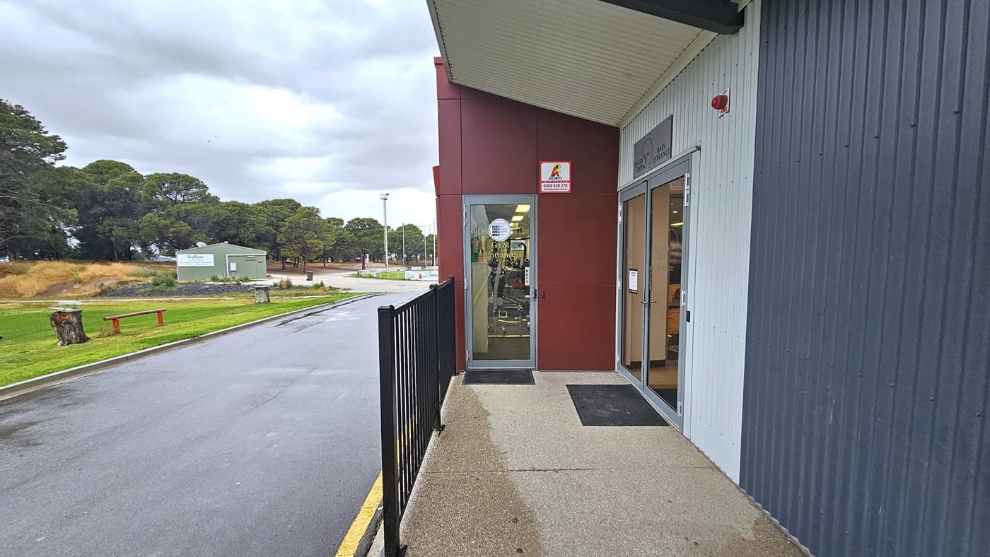 On the left is a grassy area with bench seating. A road separates the grass area from the facility building. Further ahead is a green building and a group of trees. On the right is a manual outward-opening door equipped with a non-slippery mat. Facility signage is placed on the wall. In the middle, there is a glass door that leads to the gym, with signage placed on the wall. A metal railing is positioned adjacent to the entrance.