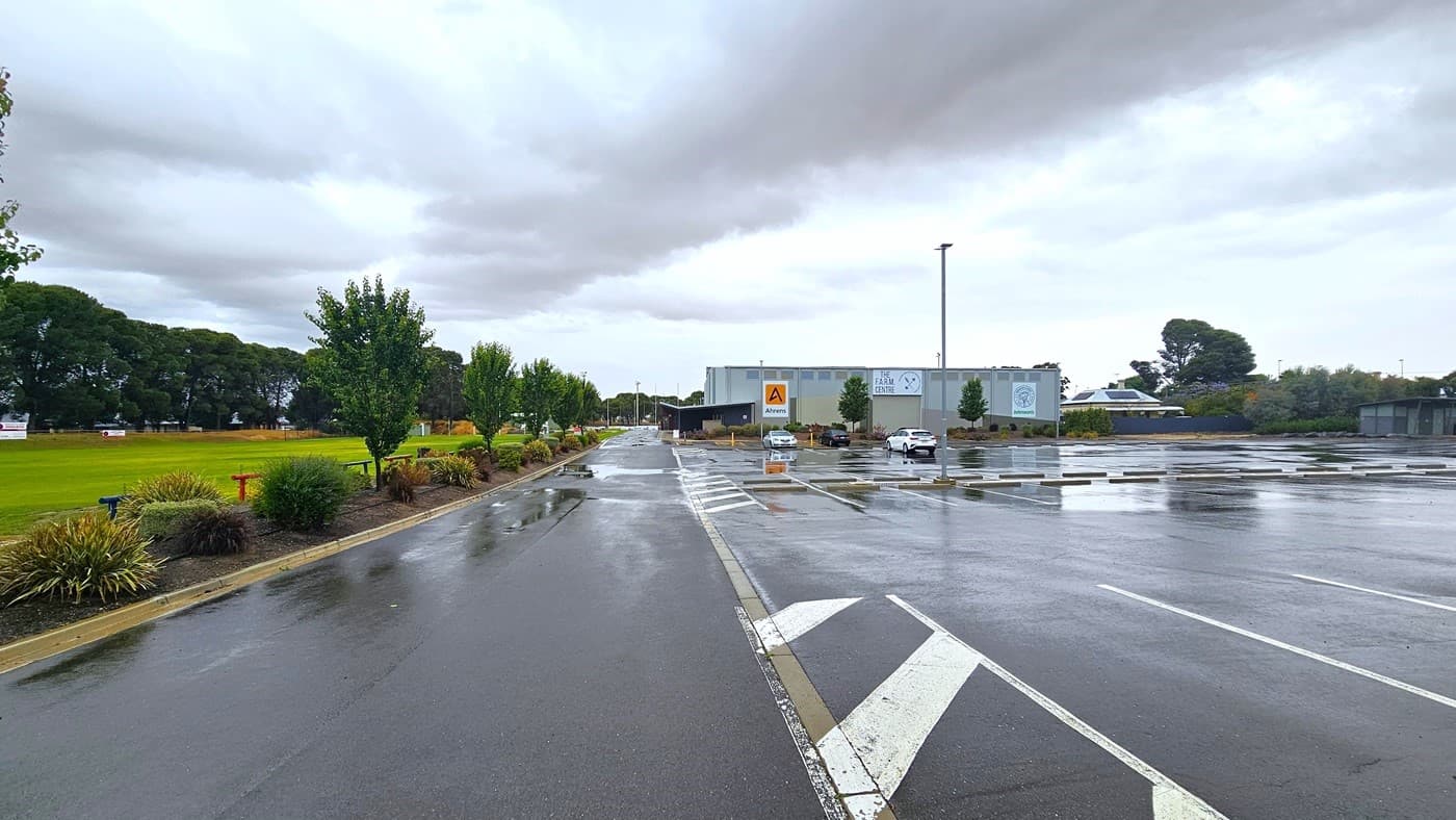 To the left is a grassy football area bordered with ornamental trees, and bench seatings are visible. On the right is an industrial building painted in grey, bearing the facility signage and some adverts. The car park is located at the side of the building, with three cars currently parked.
