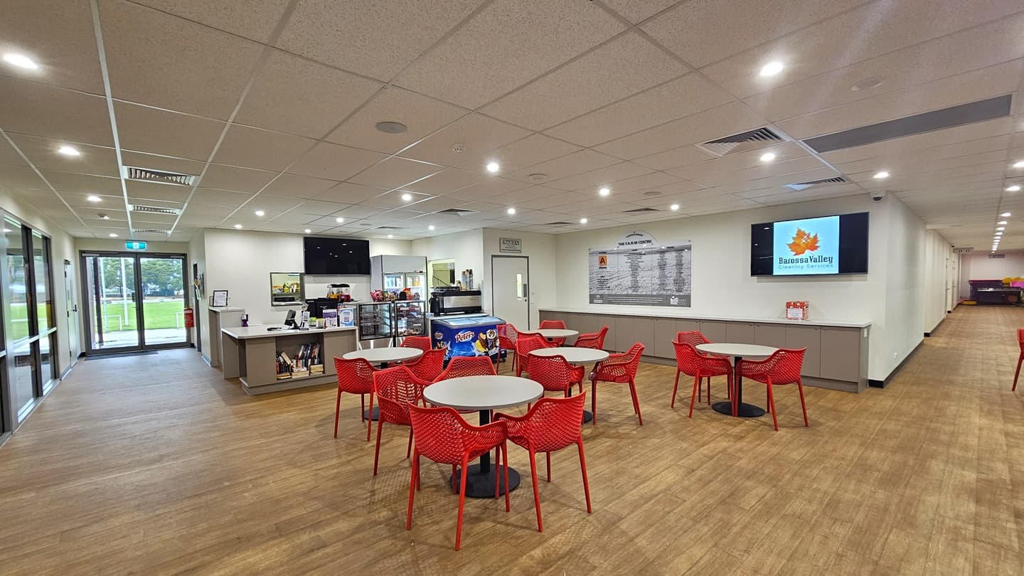 Upon entering, to the left is a floor-to-ceiling glass bearing the facility banner. A free-standing sign is also placed near the wall along with a rubbish bin. To the right is a white wall with a framed poster. Next to it is the court, also equipped with large floor-to-ceiling windows. Further in, there are red movable chairs and white tables arranged. A poster and a flat-screen TV are mounted on the middle wall. The floor is made of timber, and the ceiling is white with a porous design.