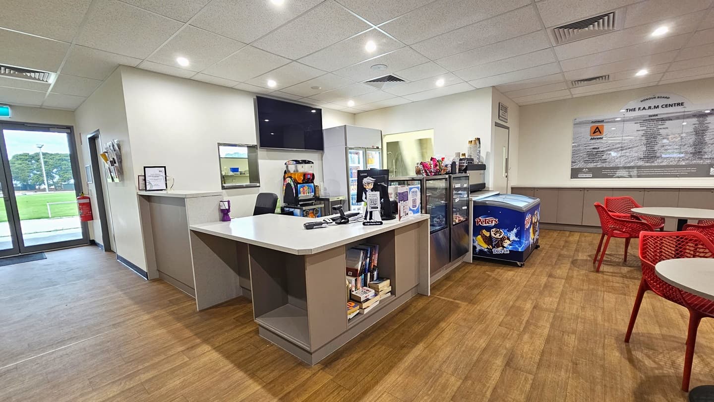 The reception desk, with a cream desk, is in the middle. A flat-screen TV is on the wall, and books are on the shelves underneath. Beside it is a display rack showcasing the cafe's items. Movable red chairs and white tables are on the right. A large poster on the wall has information. The timber floor is made of timber and the ceiling is white. To the right is a lobby leading to other facilities. On the left is a glass door with a fire extinguisher and pamphlet holder on the wall.
