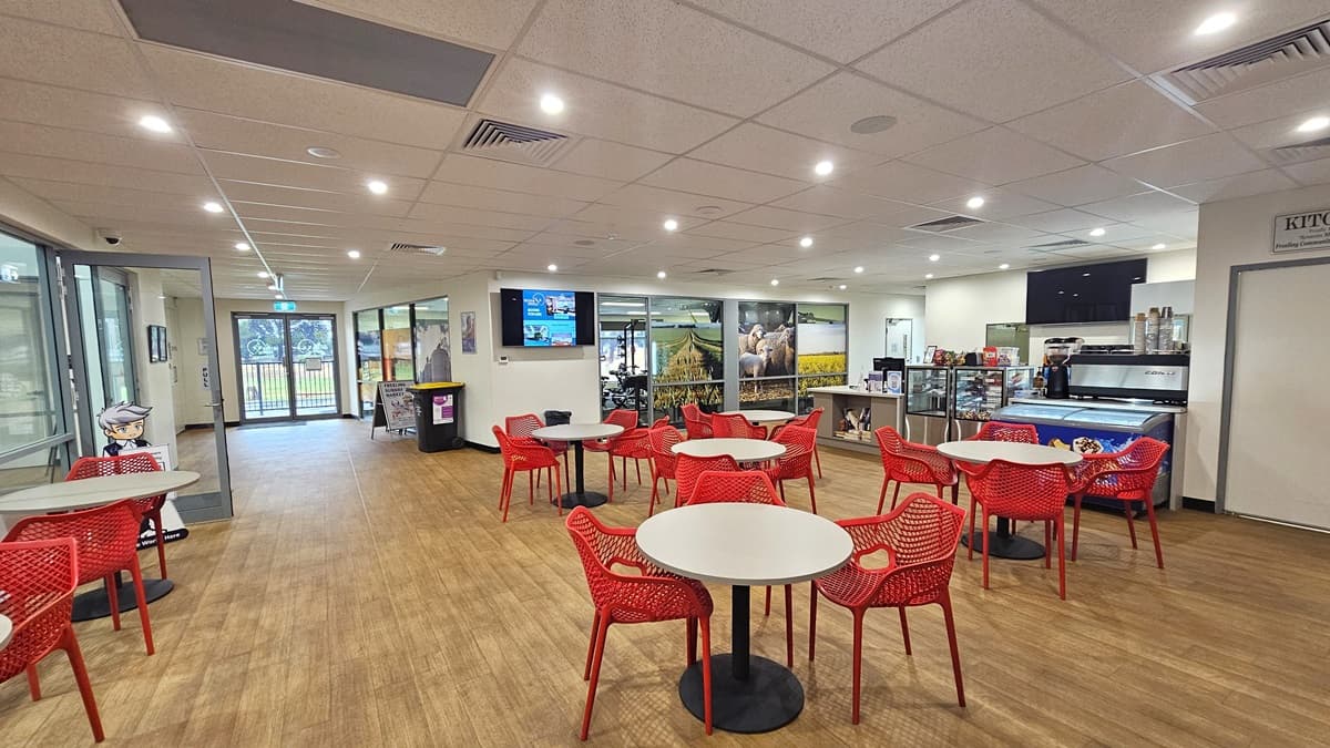 The reception desk, with a cream desk, is on the right. A flat-screen TV is on the wall, and books are on the shelves underneath. Beside it is a display rack showcasing the cafe's items. Movable red chairs and white tables are on the right. Adjacent to the reception is the gym, with a colorful glass wall and a flat-screen. To the left is the entrance to the court. Additional tables and chairs are lined up on the side. The middle wall has a rubbish bin and a free-standing sign.