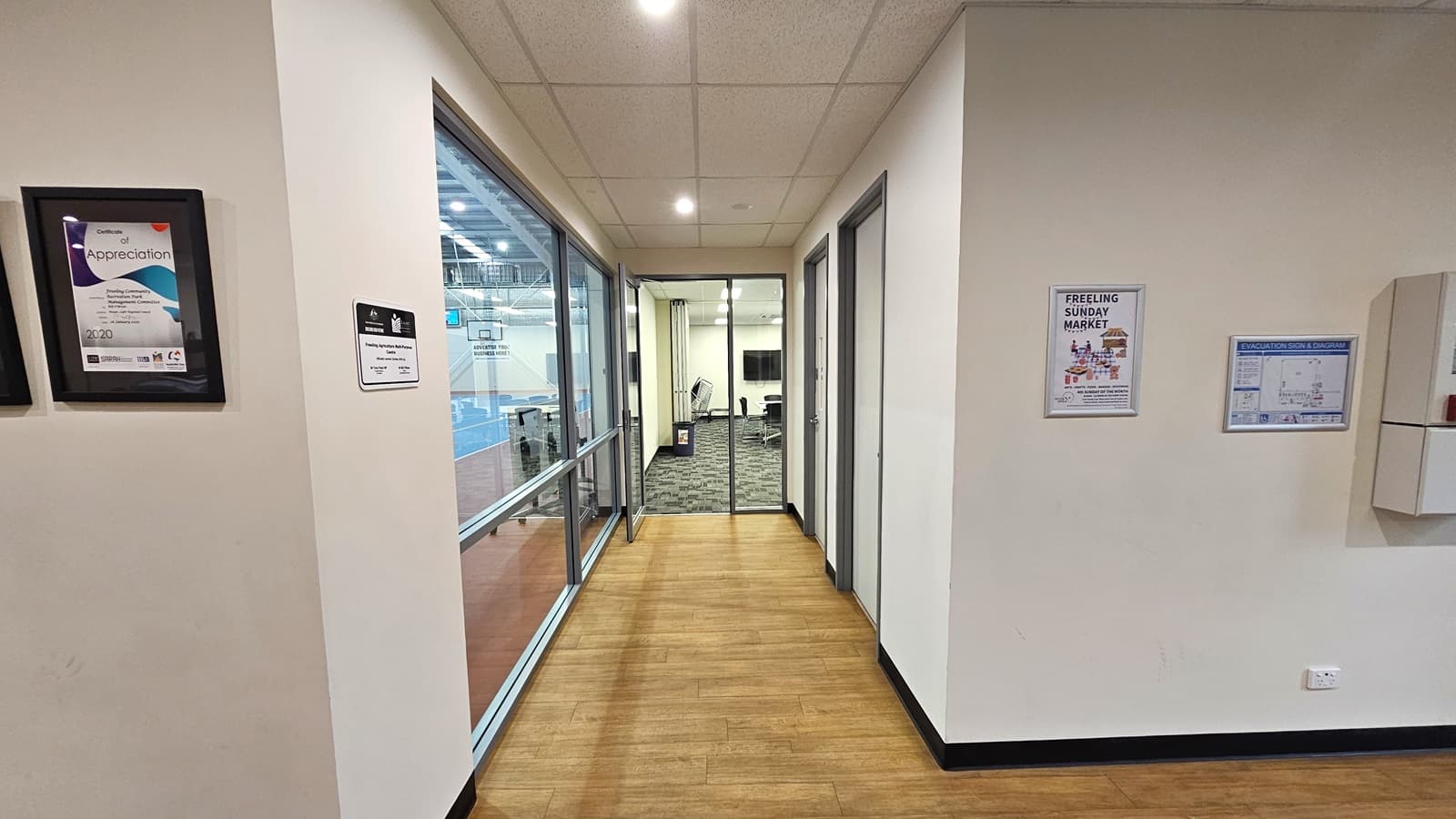 The lobby, with a timber floor, leads to the meeting room equipped with an outward-opening glass door. On the left is a floor-to-ceiling glass wall. On the right are two doors. The other wall has framed posters displaying information about the site's achievements and a venue map. The walls, including the ceiling, are white, with a porous design and circular lights.