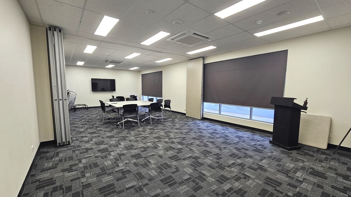 The meeting room features a spacious grey carpet with a checkered design. In the middle, there is a white conference table surrounded by black chairs. At the far end, a flatscreen TV is mounted on the wall. To the right, there are glass windows with black blinds, and a black desk is placed near the wall. On the left, an accordion steel door serves as a separator between two rooms. The ceiling is white with fluorescent lights illuminating the room.
