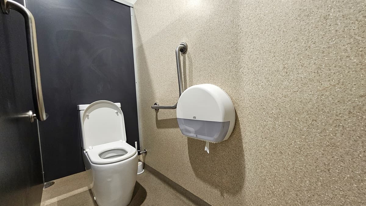 A white toilet bowl is positioned in the corner, accompanied by stainless steel handrails on both sides of the wall, along with a tissue dispenser. The left and middle walls are constructed of black granite, while the right wall is made of brown granite. The floor area is also covered in brown granite.