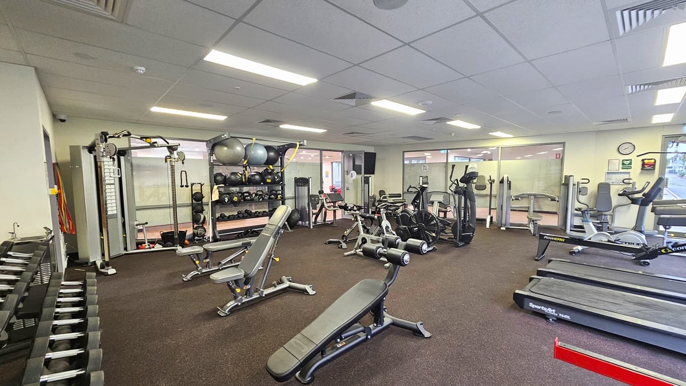 Access to the gym is through a manually operated glass door. A large flatscreen TV is mounted on the left wall near the entrance. The left wall features a floor-to-ceiling glass door adorned with a large poster of a farm landscape. Inside the gym, machines are aligned with rubber matting on the floor and a white ceiling with a porous design and centralized air conditioning. At the far end, large glass windows provide a view of the outside.