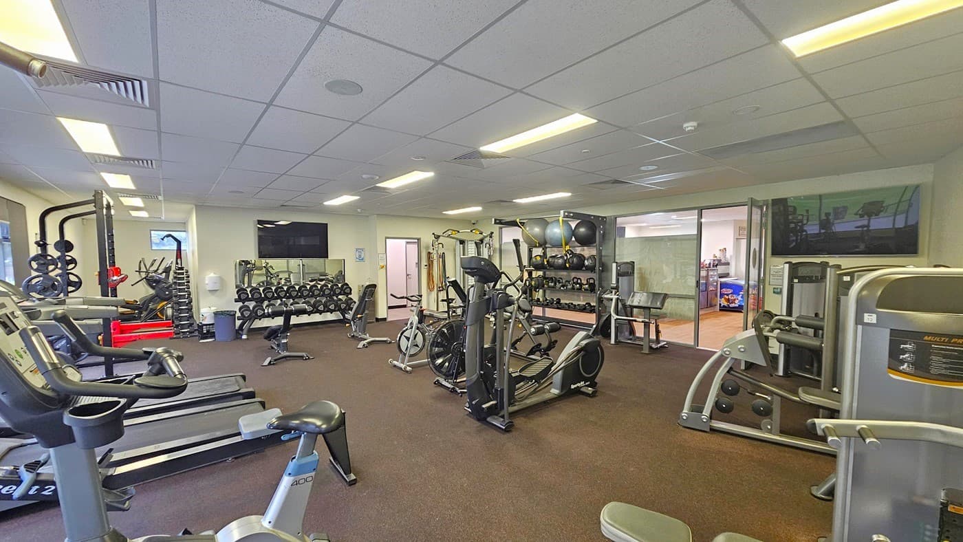 On the left, there are treadmills, stationary bikes, and Smith machines. In the middle, weights are neatly arranged in a display rack, and a flatscreen TV is mounted on the white wall. On the right, a display rack holds stability balls, medicine balls, weights, and kettlebells. Other machines are placed on the right near the door, along with a flat-screen TV. The ceiling is white with air conditioning vents.