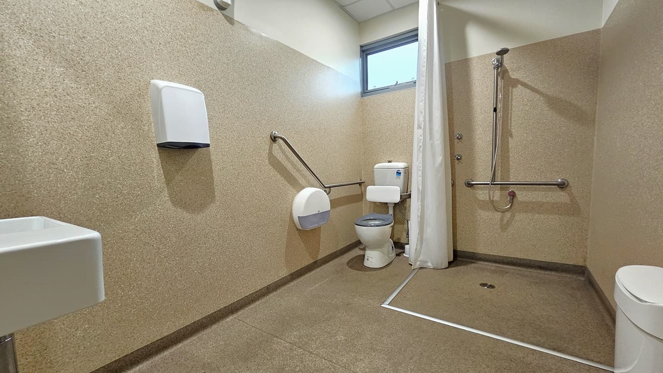 Inside the bathroom, brown granite walls enclose the space. On the left, a wash basin and hand dryer are mounted on the wall. In the corner, the toilet bowl is equipped with hand railings. A glass window is placed overhead. In the middle, a shower is mounted to the wall, separated by a white shower curtain. On the right is a rubbish bin.