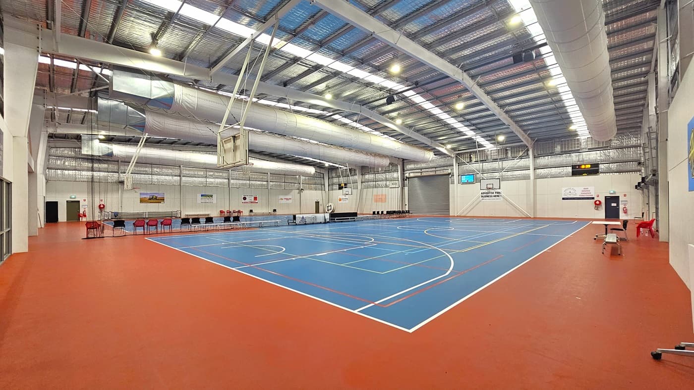 Access to the stadium is through a manual, outward-opening glass door equipped with a non-slip mat. On the right is a framed poster displaying the site's achievements. To the left is a table with a red plastic chair. Inside the stadium, a large and open space, the size of two basketball courts marked for various ball games. The ceiling is adorned with exposed white beams, reflective silver insulation, and a mixture of natural skylights and spotlights.