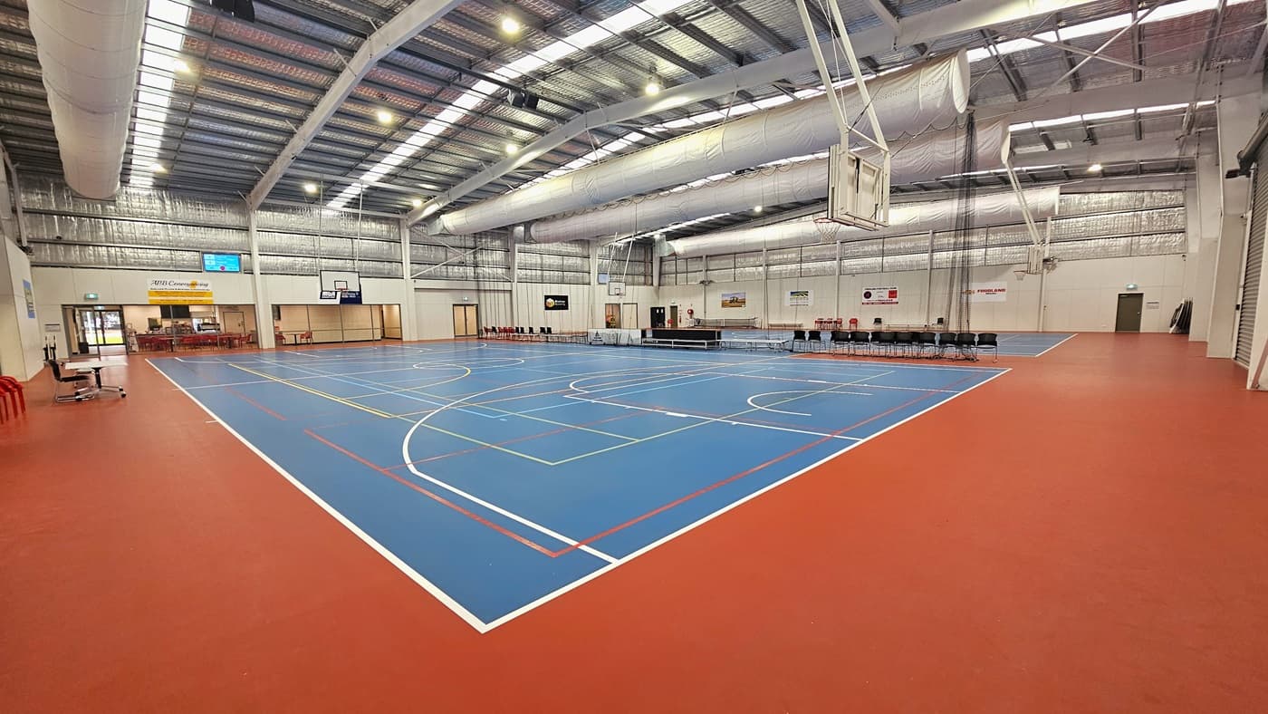 The solid floor exhibits a combination of blue and red, with markings for various ball games. The ceiling showcases exposed white beams, air conditioning vents, reflective silver insulation, and a blend of natural skylights and spotlights. In the middle, chairs and tables are arranged to distinguish the two courts. Digital screens are affixed to the left walls, and tables and chairs are positioned on the left. Basketball boards are installed on both sides.
