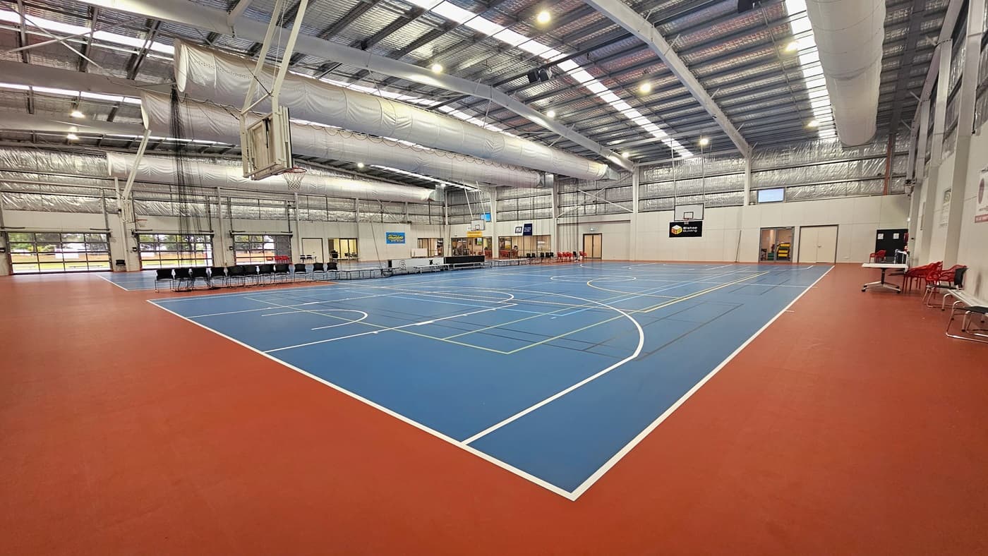 The floor features a blend of blue and red with markings for various ball games. The ceiling displays exposed white beams, air conditioning vents, reflective silver insulation, and a mix of natural skylights and spotlights. In the middle, chairs and tables are arranged to separate the two courts. Digital screens are mounted on the middle walls. Basketball boards are positioned on both sides. On the left, tables and chairs are placed near the white wall.