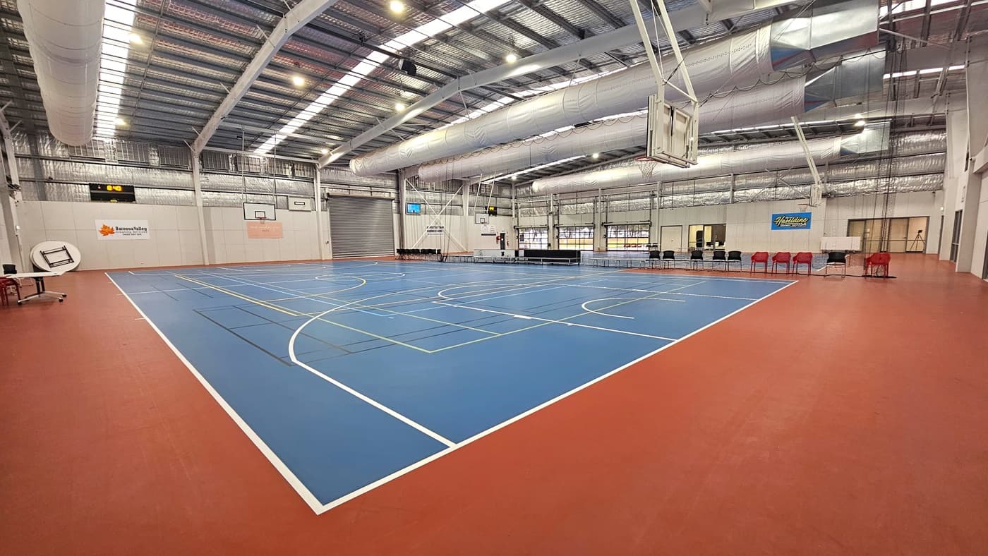 The solid floor exhibits a combination of blue and red, with markings for various ball games. The ceiling showcases exposed white beams, air conditioning vents, reflective silver insulation, and a blend of natural skylights and spotlights. In the middle, chairs and tables are arranged to distinguish the two courts. Digital screens are affixed to the middle walls. Basketball boards are installed on both sides.In the left, tables and chairs are positioned near the white wall.