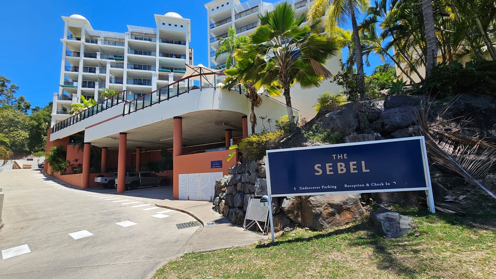 A multi-storey building with white balconies and a flat roof is on the left. Below, there is a covered parking area supported by orange columns. A driveway leads up to the building. On the right, a sign reads "The Sebel" with directions for parking and reception. The area is landscaped with palm trees and rocks, and there is a grassy patch in the foreground.