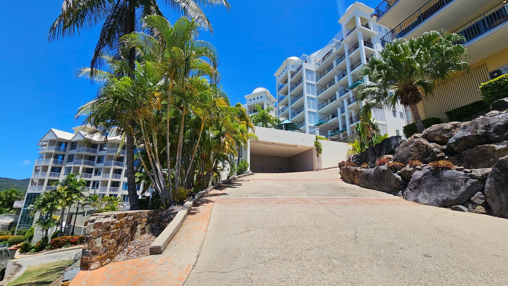 A sloped driveway leads up to a large, white multi-storey building with balconies on the right. On the left, there are tall palm trees and another similar building in the background. The foreground features a stone wall and landscaped rocks with small plants. The sky is clear and blue.