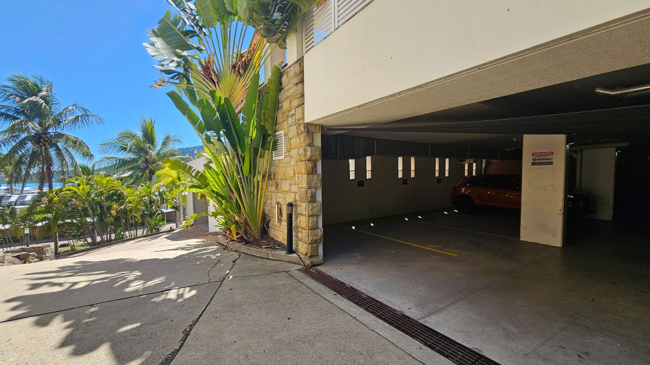 On the left, there are tall palm trees and lush greenery lining a sloped driveway. In the middle, a stone wall with large tropical plants is adjacent to an open garage. On the right, the garage interior is visible, with a car parked inside. The floor is concrete, and the ceiling is flat with fluorescent lights.