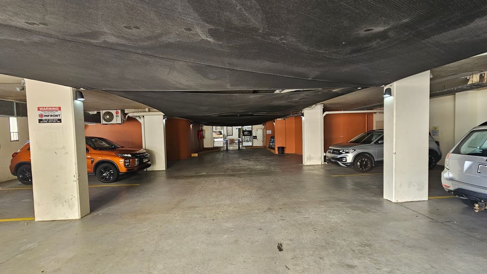 A parking garage with a concrete floor and a dark, textured ceiling. On the left, an orange car is parked next to a column with a warning sign. On the right, a silver car is partially visible. The background shows a small office area with a desk and various signs. The walls are painted in a mix of orange and beige.