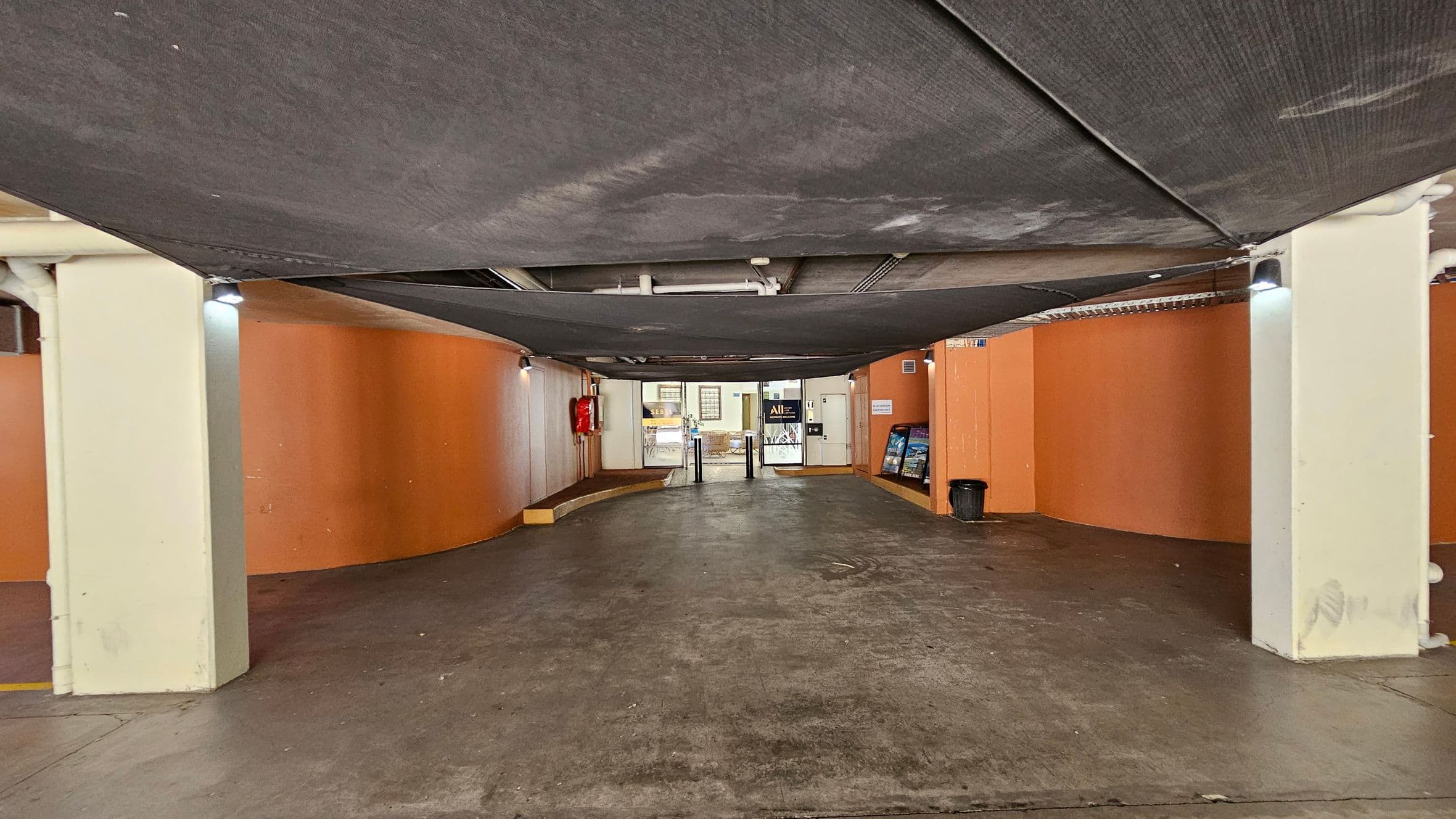 A covered parking area with a concrete floor and orange walls. The ceiling is draped with dark fabric panels. On the left, there's a fire extinguisher mounted on the wall. In the centre, a glass entrance with turnstiles leads to another area. On the right, a black bin is placed against the wall. The lighting is provided by small fixtures attached to the walls.
