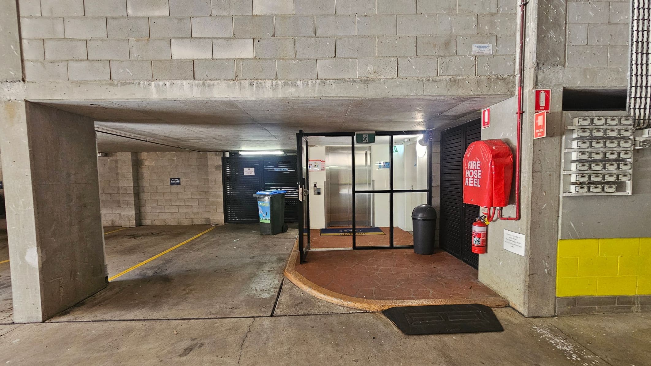 A parking garage with concrete walls and ceiling. On the right, a red fire hose reel and extinguisher are mounted on the wall. A series of electrical boxes are visible below a yellow stripe. In the centre, a glass door leads to an elevator lobby with a mat on the floor. A rubbish bin and a recycling bin are placed outside the door. The floor is concrete with yellow parking lines. The ceiling is low and made of concrete.