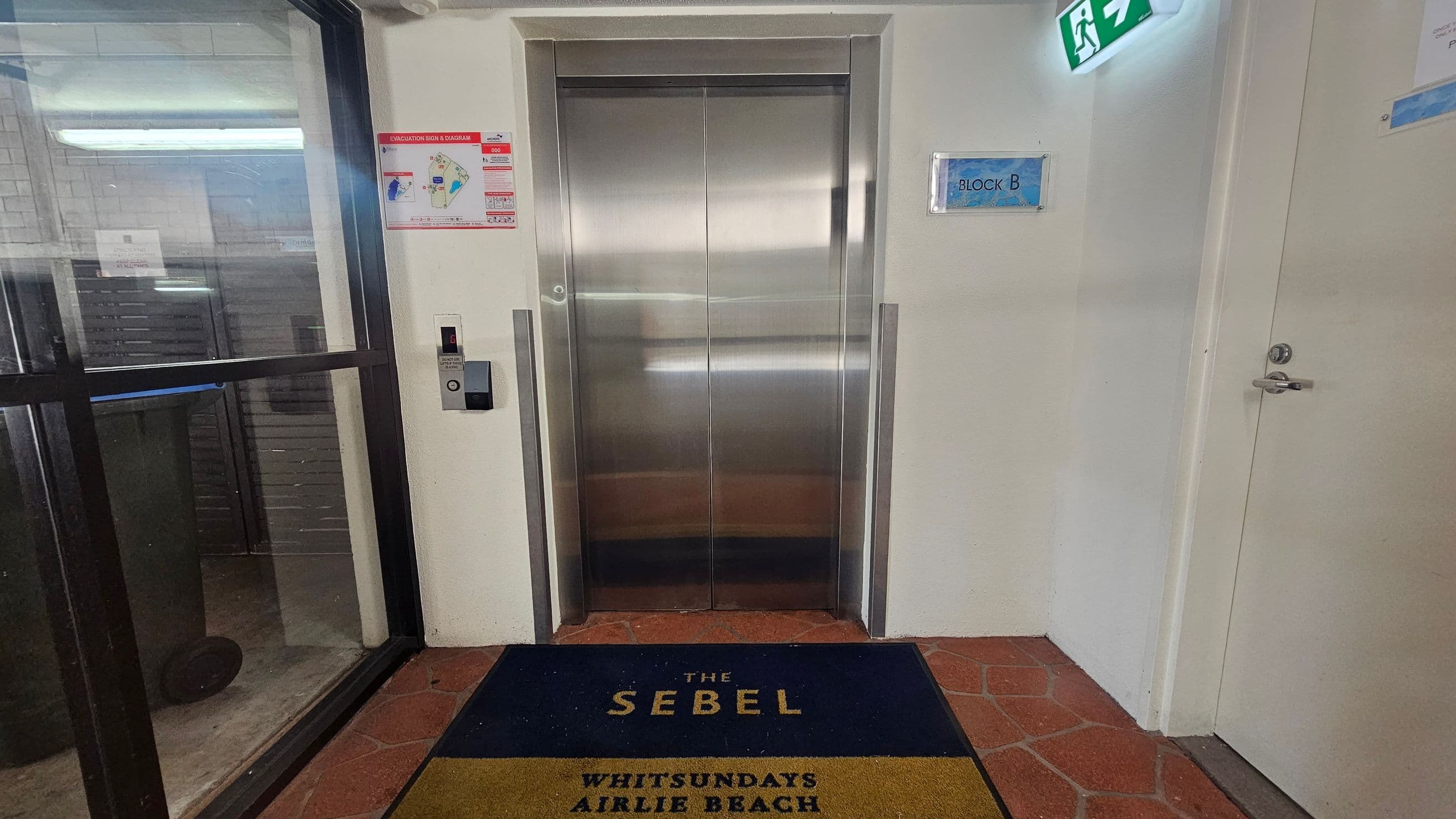 A lift with closed metallic doors is centred on a wall. To the left, there's a glass door with a reflection of fluorescent lights. A lift call button is mounted on the wall next to the lift. On the right, a door with a handle is visible, along with a sign reading "BLOCK B" and an illuminated exit sign above. The floor is covered with a mat that reads "THE SEBEL WHITSUNDAYS AIRLIE BEACH" on a tiled surface. An evacuation sign is posted on the wall near the lift.