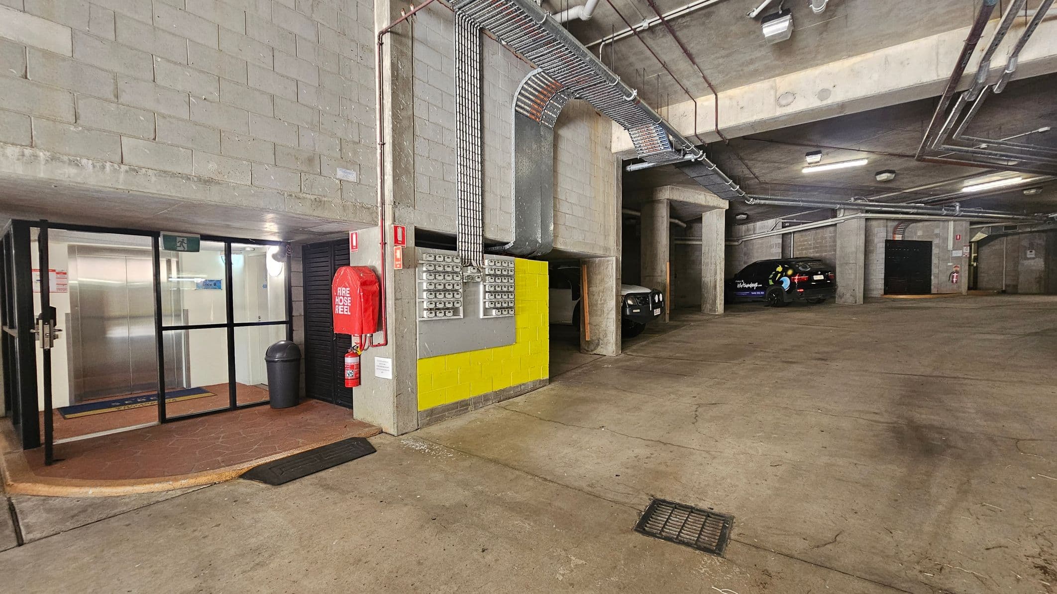Underground car park with concrete walls and ceiling. On the left, a glass door entrance with a black frame leads to a hallway with a visible lift. A red fire hose reel and extinguisher are mounted on the wall next to a yellow panel with electrical boxes. The floor is concrete with a drain cover. On the right, two parked cars are partially visible in separate bays. Overhead, exposed pipes and cables run along the ceiling.