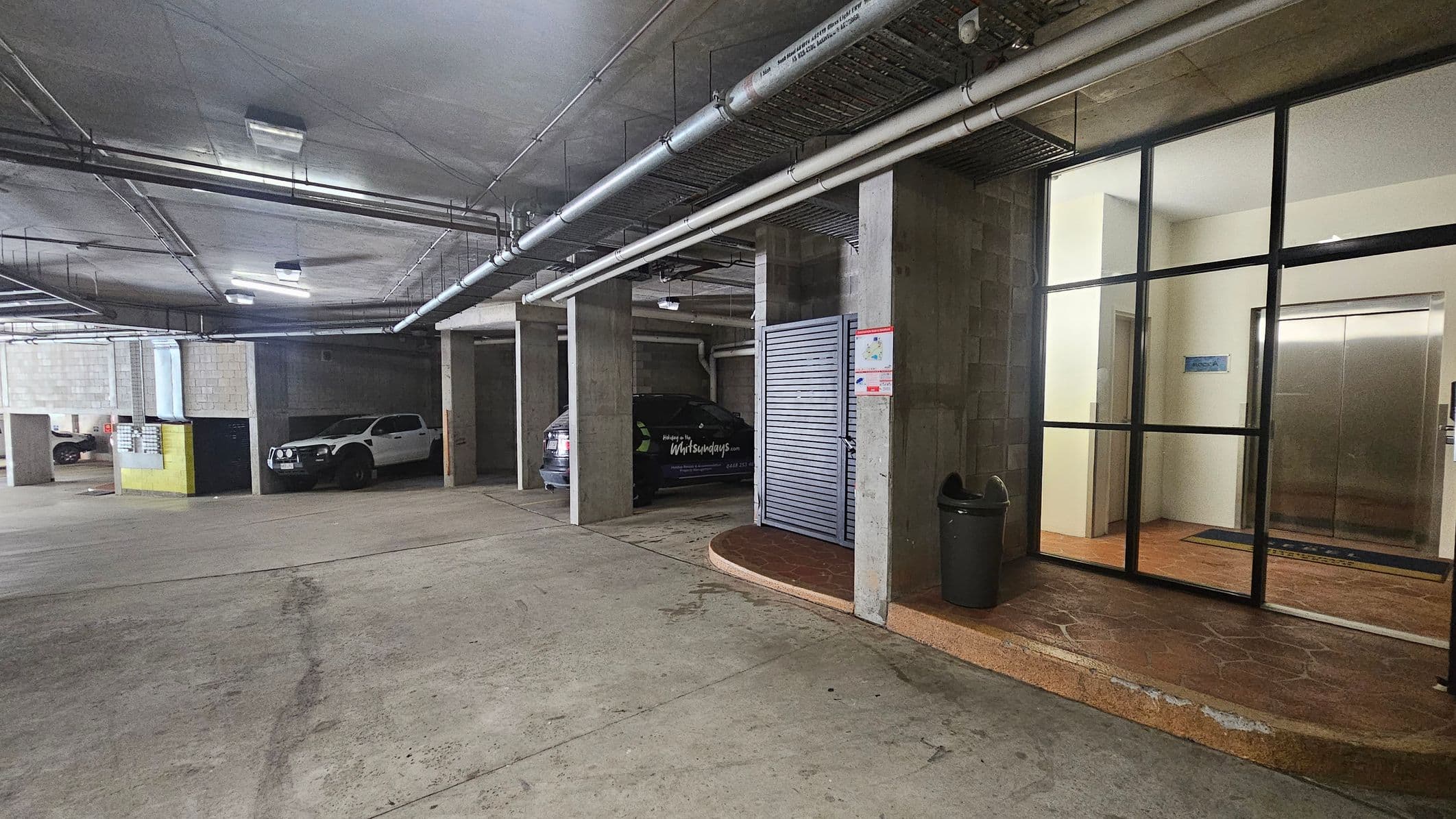 Underground car park with a concrete floor and ceiling. On the left, two vehicles are parked in separate bays, one white and one dark. In the centre, a grey metal door is next to a black car. On the right, a glass wall reveals an interior space with an elevator and a wooden floor. Above, exposed pipes and lighting fixtures run across the ceiling. A black bin is near the entrance.