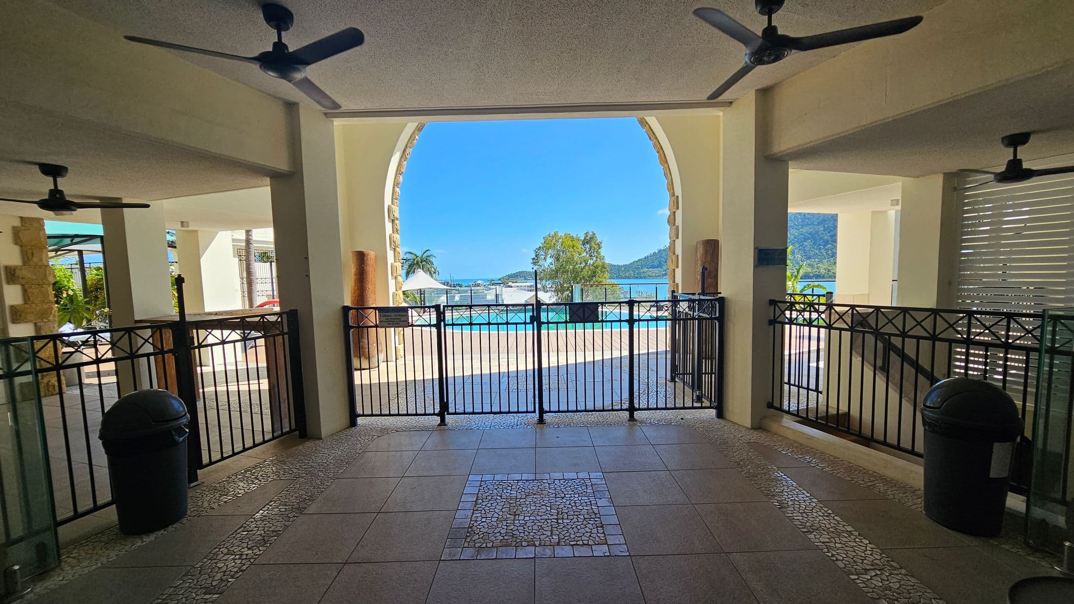 A view through an archway with a black metal gate leading to an outdoor pool area. The foreground has a tiled floor with a mosaic pattern. Ceiling fans are visible above. On the left and right, there are black bins and stone pillars. Beyond the gate, a pool is visible with a backdrop of trees, a palm tree, and distant hills under a clear blue sky.