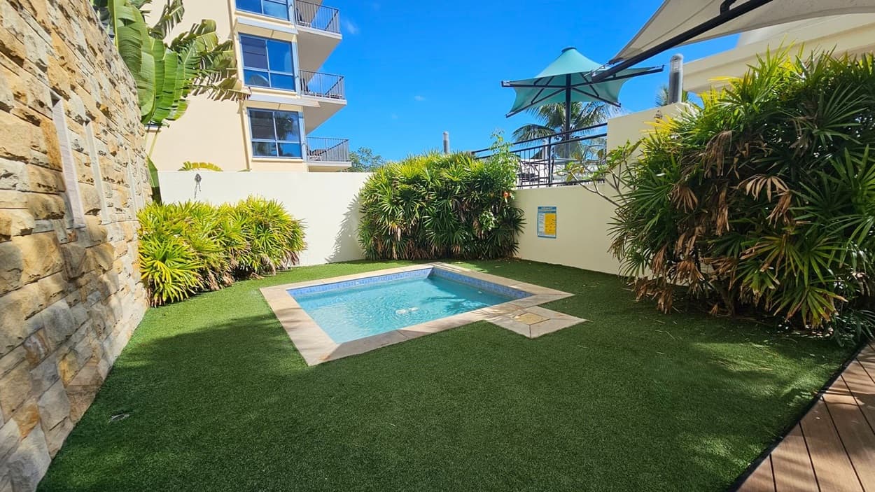 A small square pool is surrounded by artificial grass. On the left, there's a stone wall with tropical plants. In the background, a multi-storey building with balconies is visible. On the right, a white wall with dense green foliage and a sign is present. Above, a large umbrella provides shade.