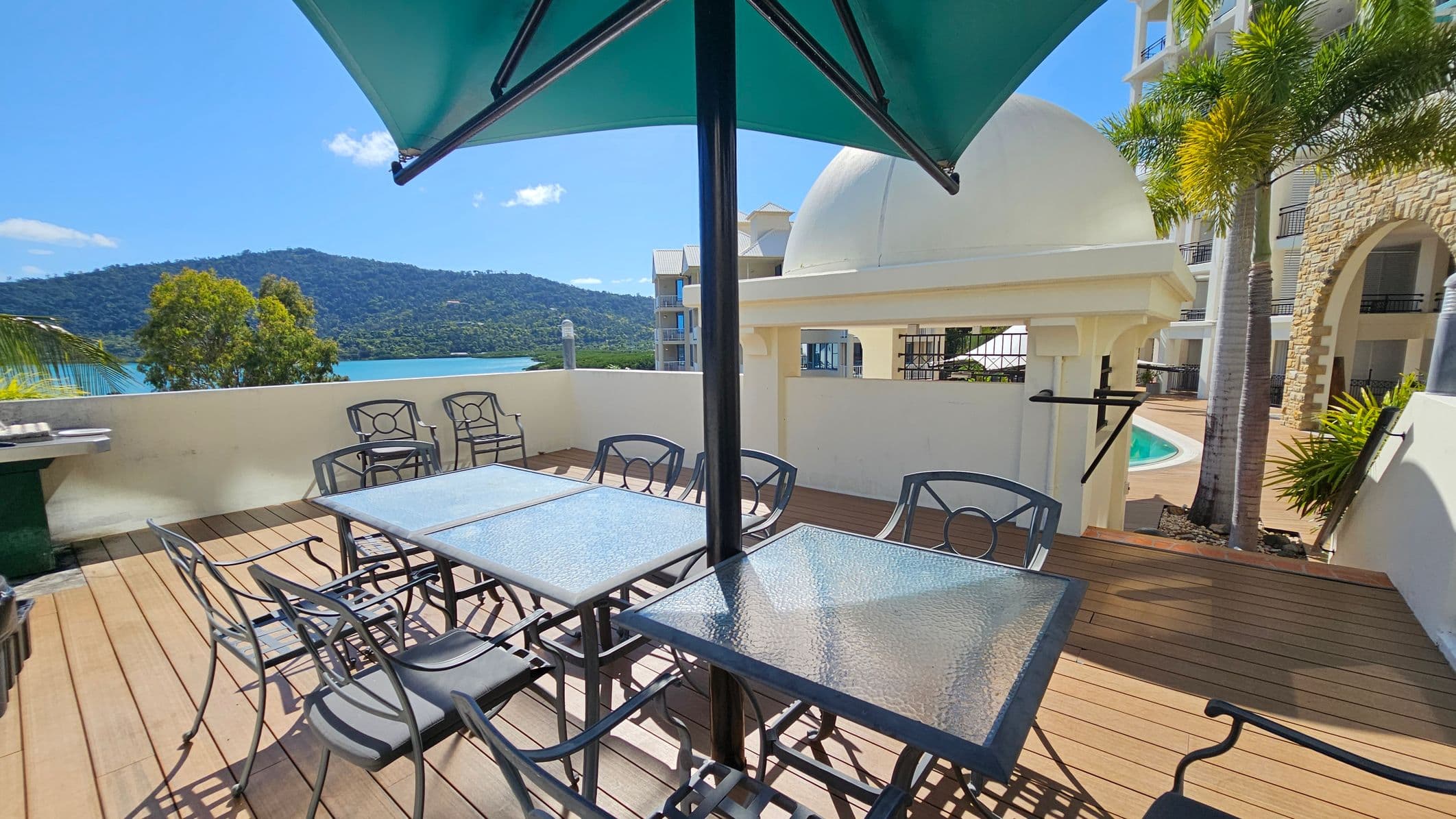 Outdoor terrace with wooden decking and glass-topped tables surrounded by metal chairs. A large green umbrella provides shade. In the background, a white domed structure and a swimming pool are visible, along with a stone building facade. To the left, a view of a lush green hill and a body of water can be seen over a low wall. Palm trees are present on the right side.