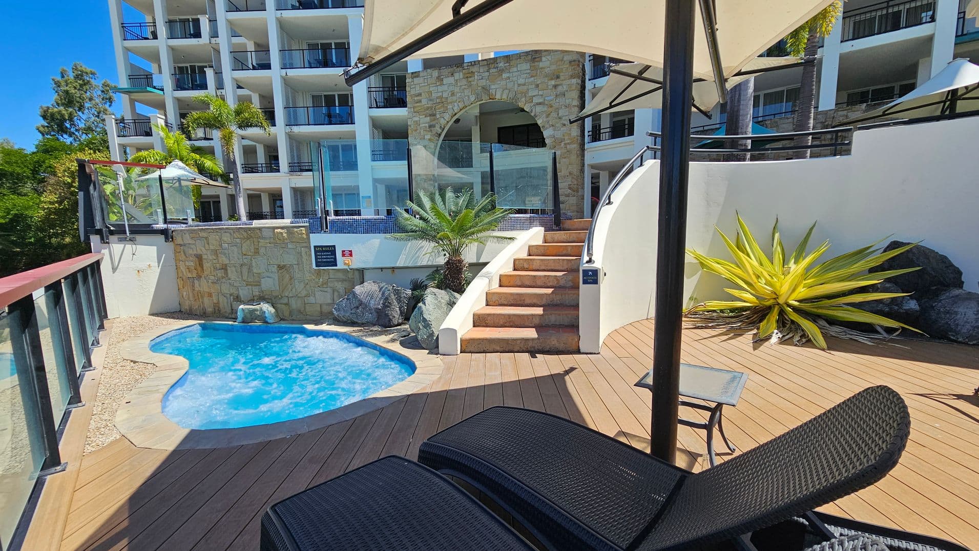 A small, curvy pool is on the left, surrounded by a stone patio. A stone wall with a glass barrier and a staircase leads up to a higher level. On the right, a large, spiky plant is next to some rocks on a wooden deck. A black wicker sun lounger and a small table are in the foreground. In the background, a multi-storey building with balconies and palm trees is visible. An umbrella provides shade over part of the scene.